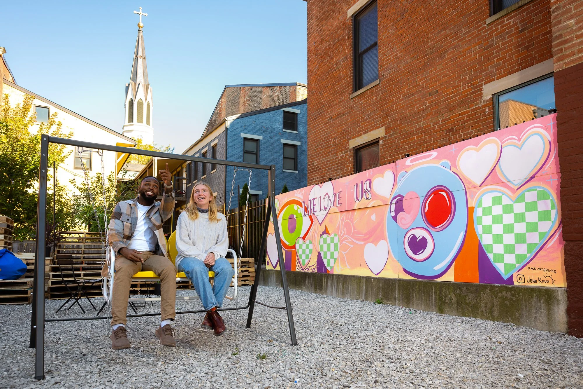 Two people sitting on a swing in an outdoor urban space with colorful wall art that says "LOVE US" and features hearts and other colorful designs.