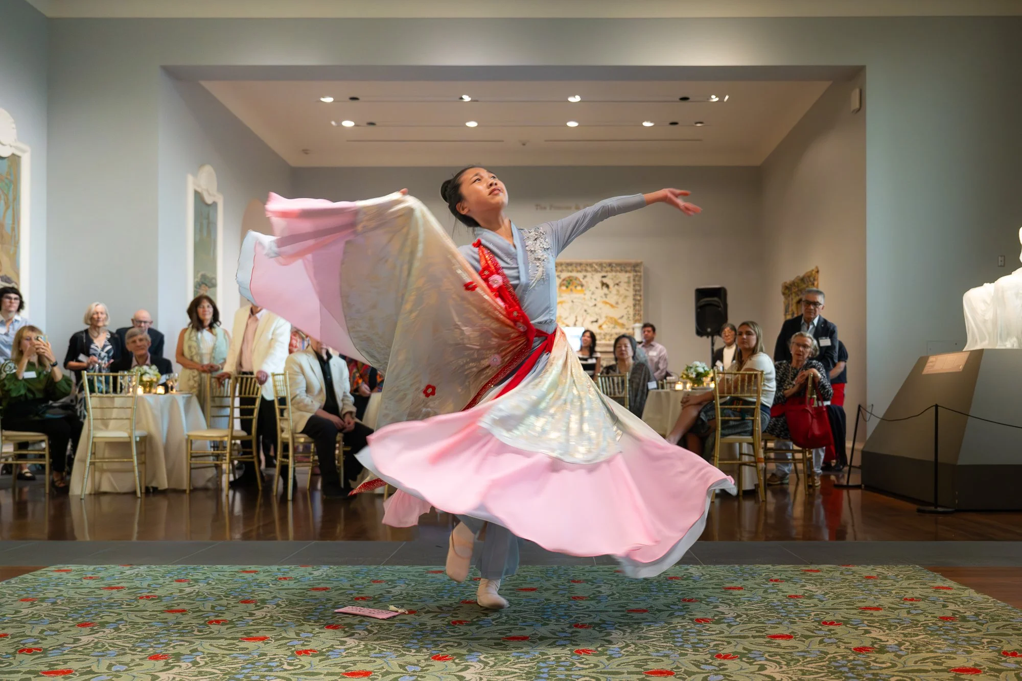 A woman in traditional Korean hanbok dancing in an art museum, with an audience seated and standing around her observing the performance.