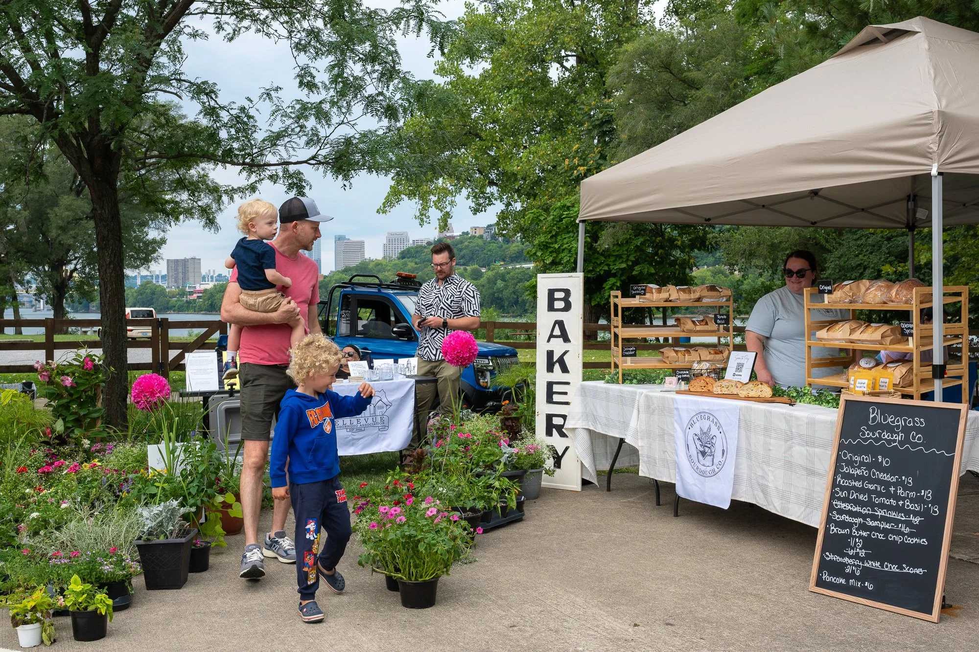 An outdoor bakery stand with baked goods on display, a woman behind the table, a sign reading 'Bakery,' a chalkboard menu, and customers browsing and shopping among potted plants and flowers on a sidewalk near a river with cityscape in the background