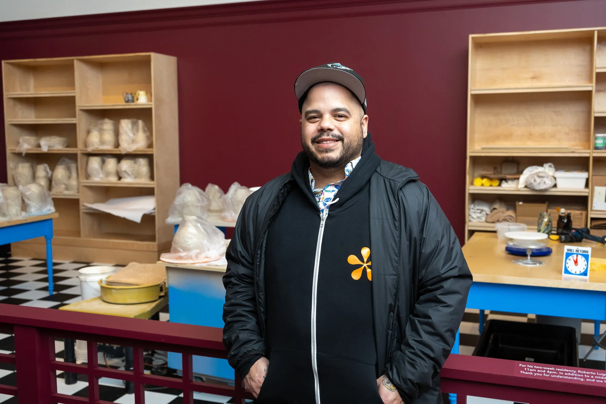 A man smiling, wearing a black jacket with an orange flower logo, standing in a room with wooden shelves and a checkered black-and-white floor.