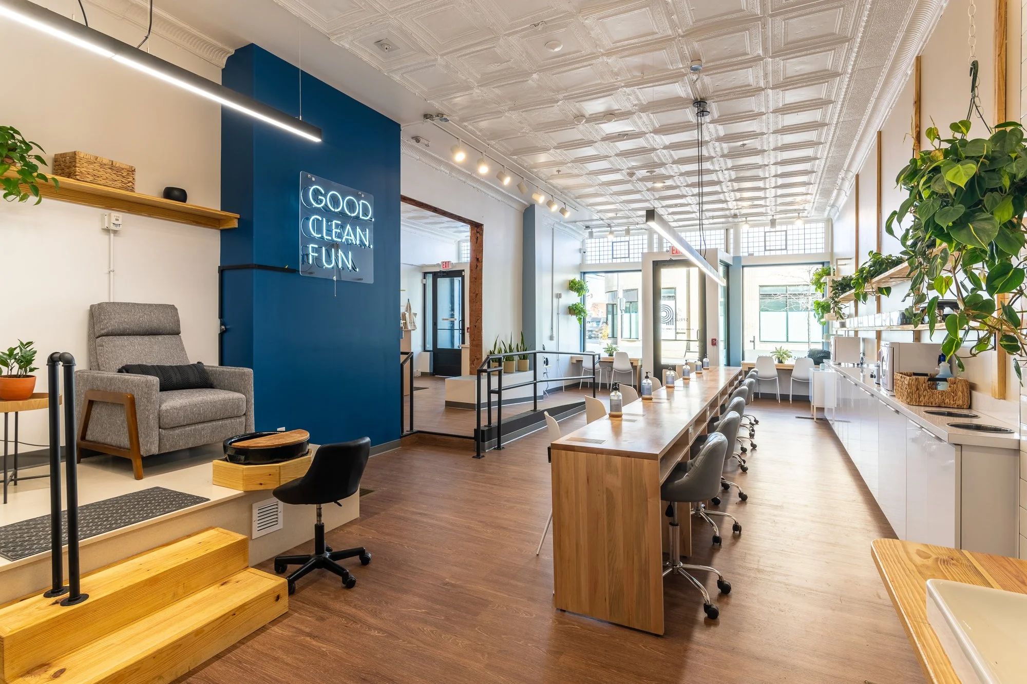 Bright, modern workspace with a long wooden table, white chairs, and plants. A navy blue accent wall features a neon sign reading 'Good Clean Fun.' The area has large windows, a white ceiling with decorative tiles, and a kitchenette with a sink and s