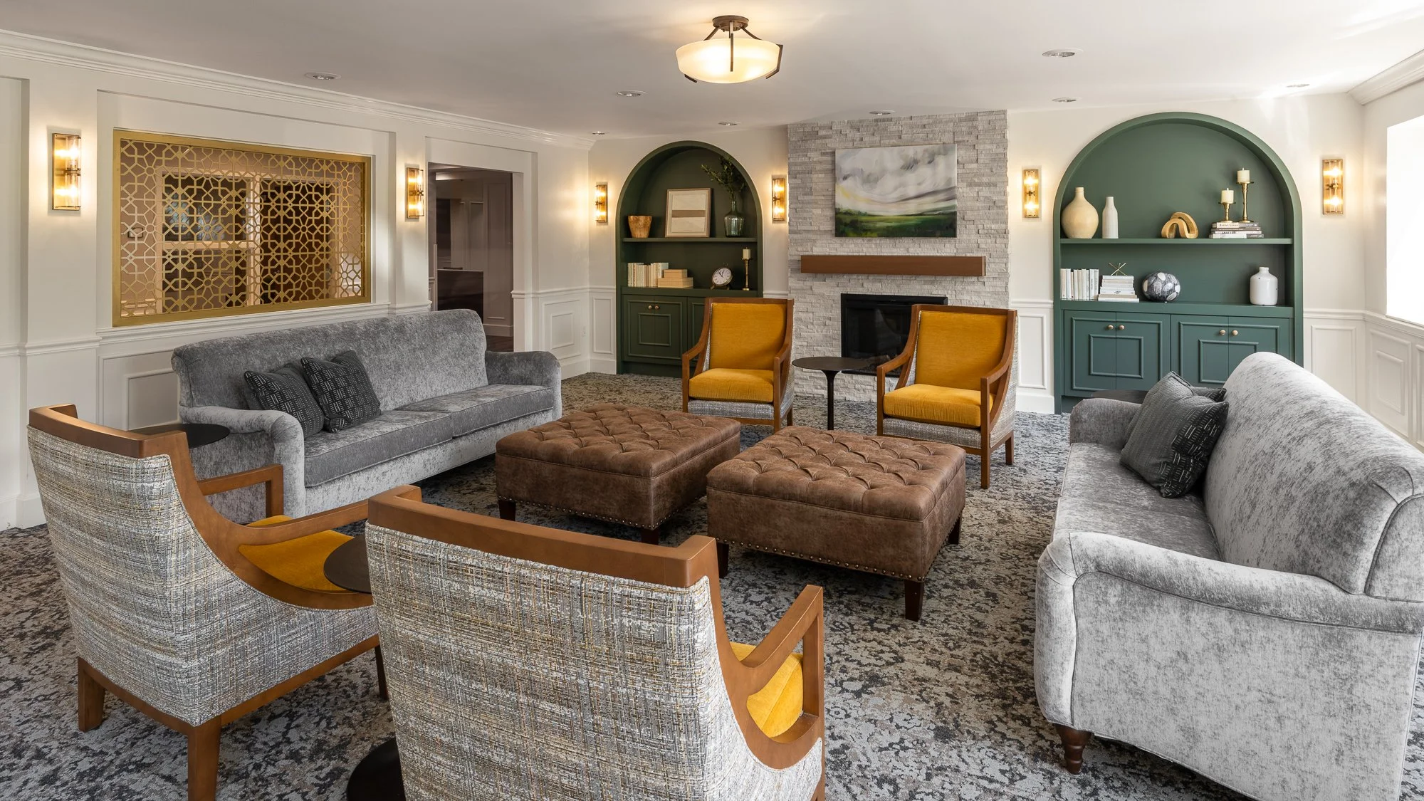 Living room with two gray sofas, two armchairs with yellow cushions, two brown tufted ottomans, a fireplace, built-in shelves with decor, and a patterned carpet.