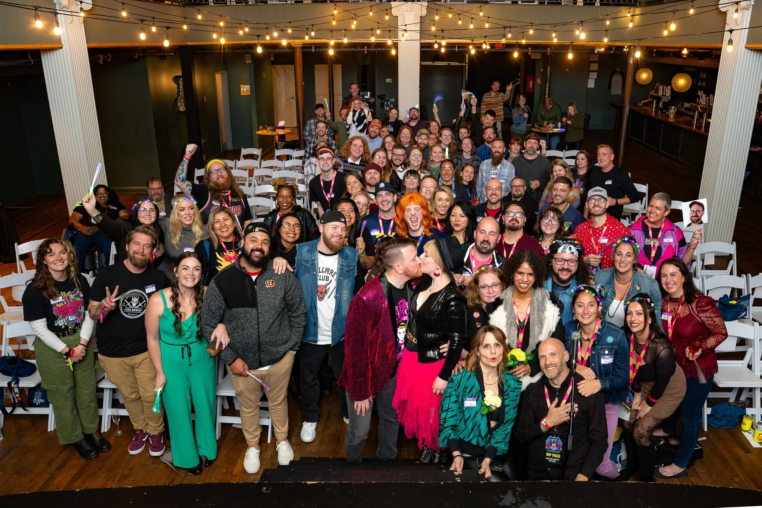 A large group of diverse people gathered indoors for a celebration or event, with some individuals holding signs and posing for a photo, and two people kissing in the front. The room has string lights hanging from the ceiling, and there are chairs an