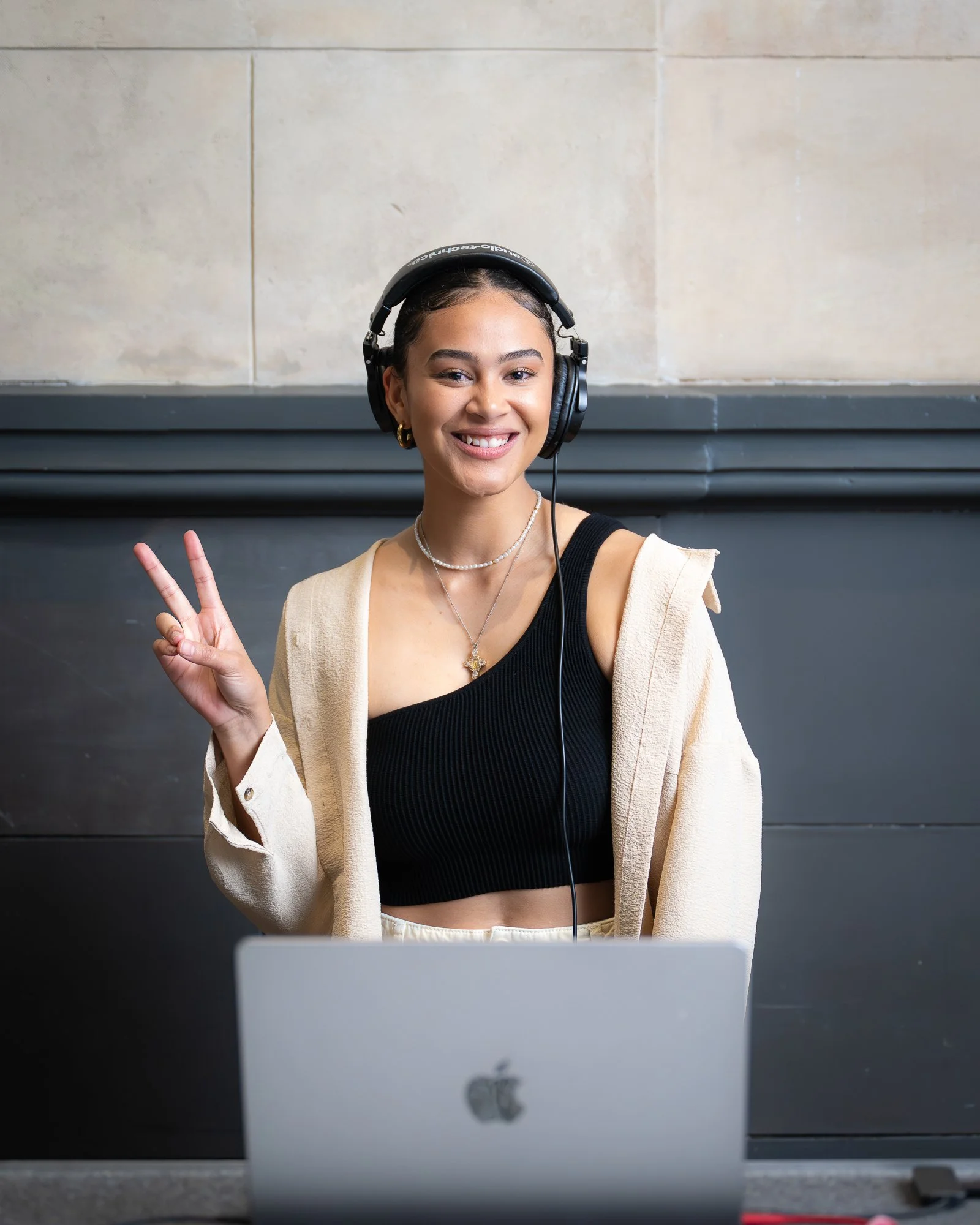 A young woman wearing headphones, smiling, and making a peace sign with her hand, seated in front of a laptop.
