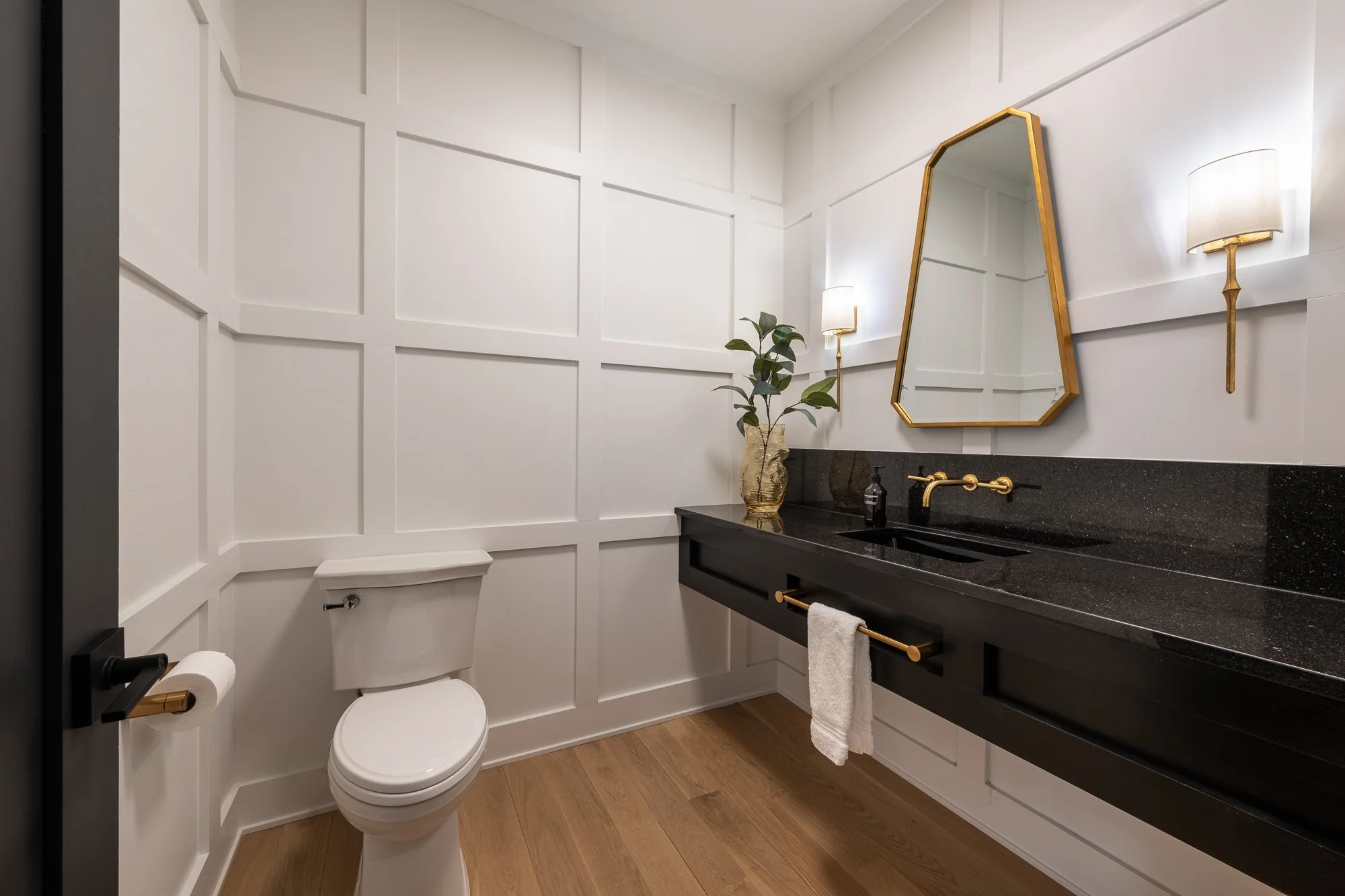 Modern bathroom with white paneled walls, a black vanity with gold fixtures, a mirror, wall-mounted lights, and a potted plant.