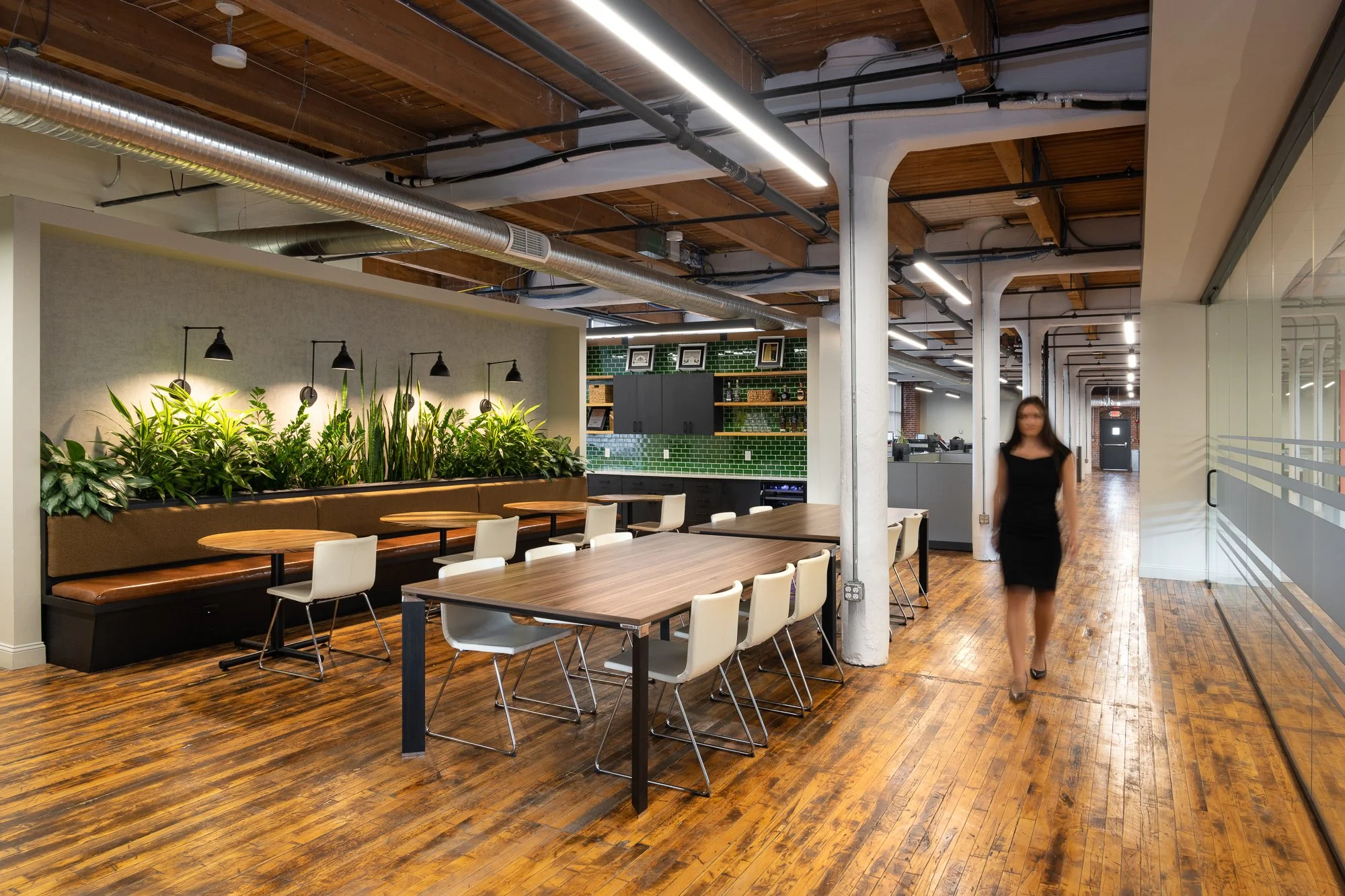 Modern office break room with wooden flooring, plants, tables, and white chairs, featuring an open ceiling with visible ductwork and industrial design elements.