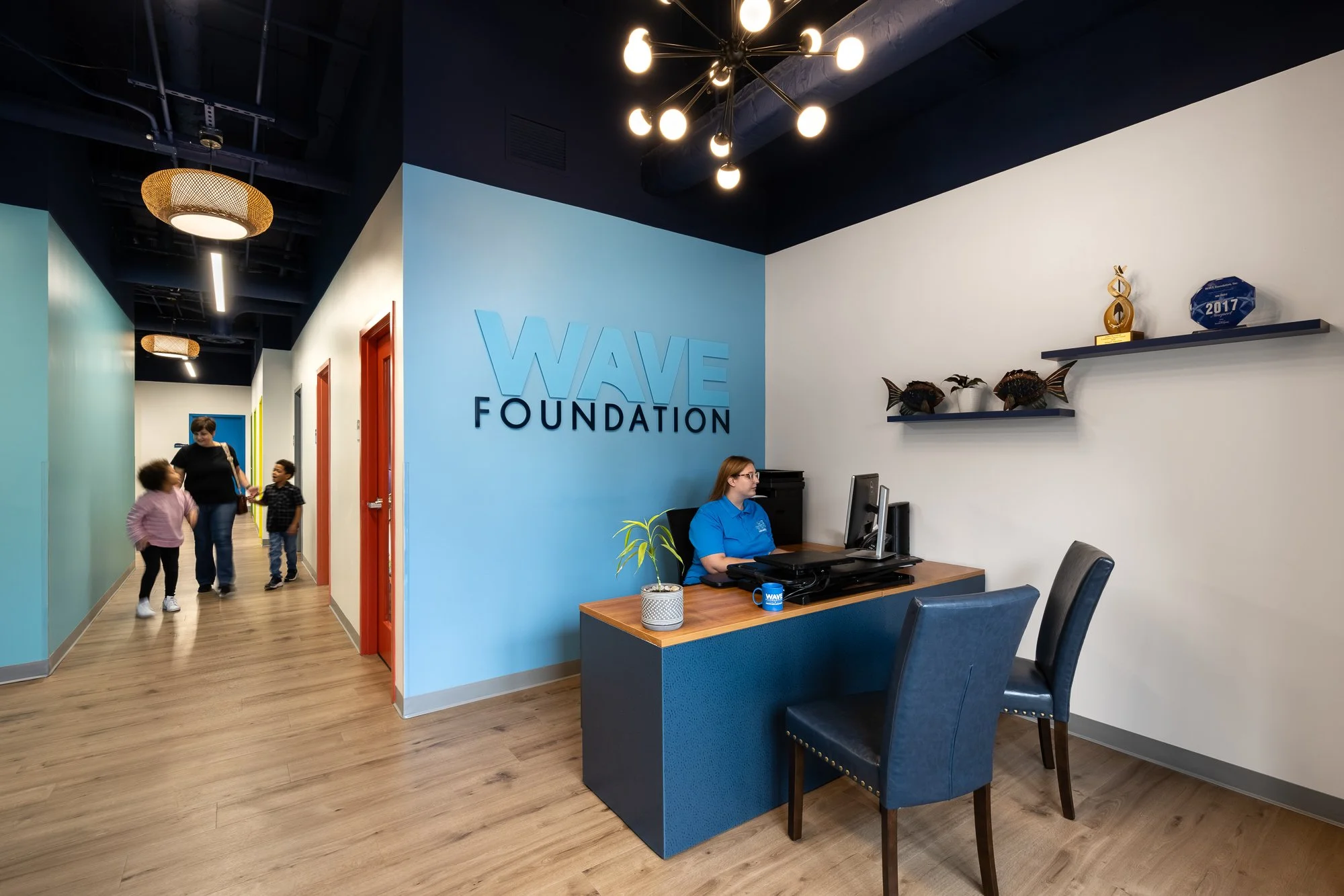 Reception area with a woman at a desk, wall sign reading 'WAVE FOUNDATION'; hallway with children and an adult, colorful walls, and decorative ceiling lights.