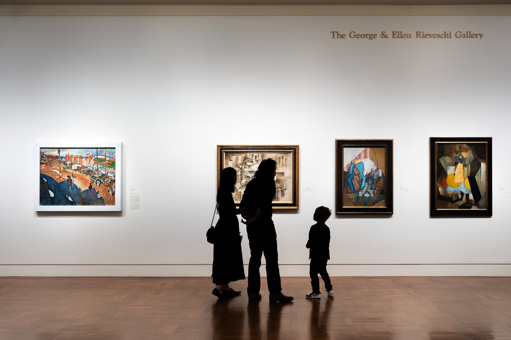 Silhouettes of a woman, man, and child standing in front of framed paintings in an art gallery, with the gallery's name, 'The George & Ellen Rieveschl Gallery,' on the wall.
