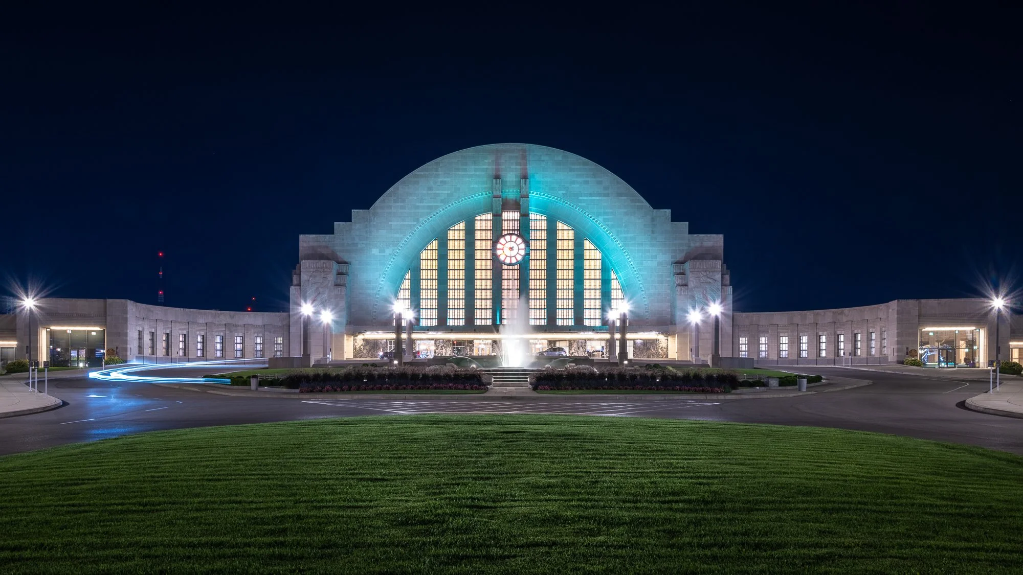 Night view of a large, historic train station with illuminated windows and a central fountain, surrounded by a well-maintained lawn and circular driveway.