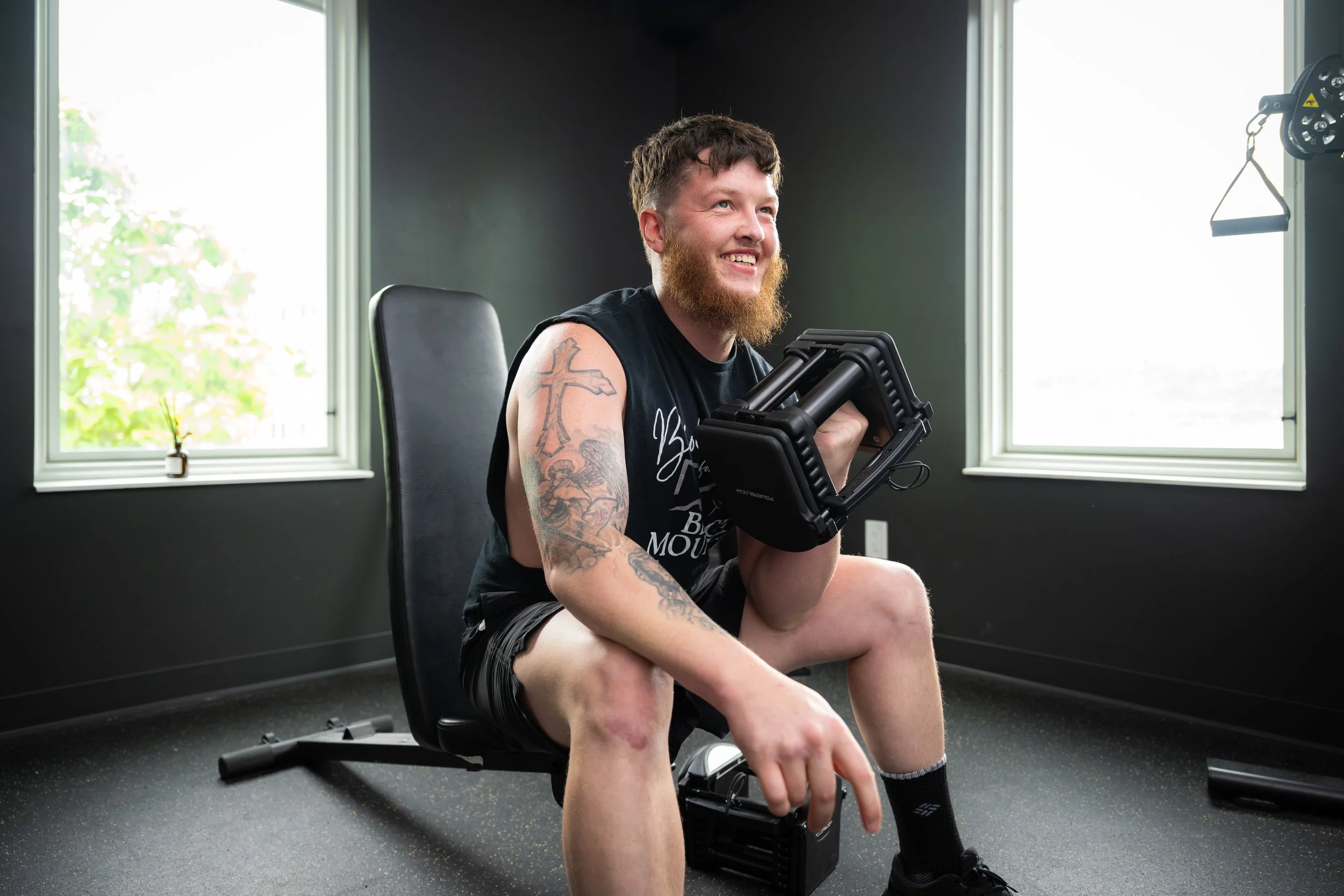 A man with a beard and tattoos sitting on a workout bench holding a small exercise machine, smiling inside a gym with black walls and two windows.