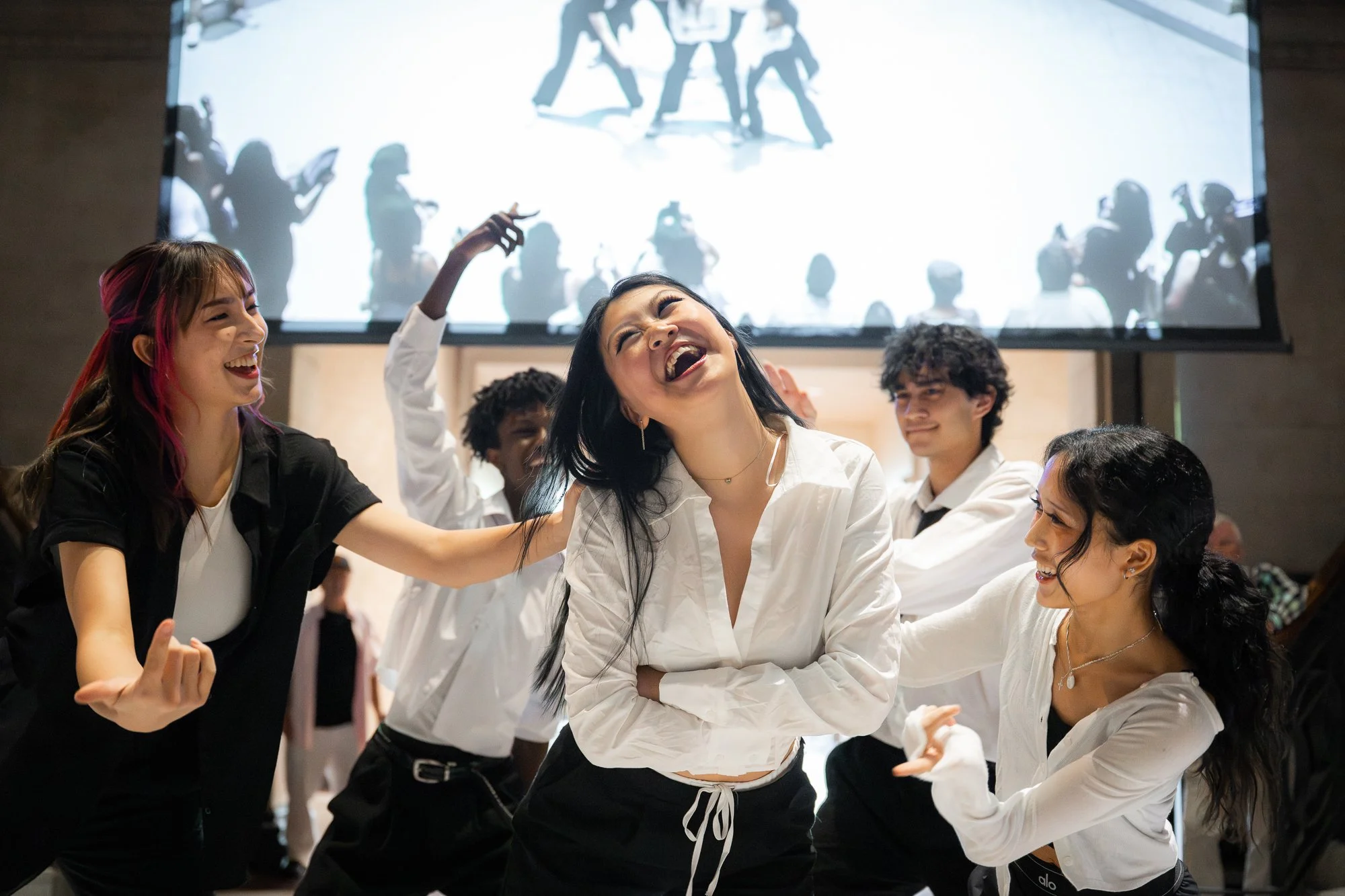 A group of diverse young people dancing and laughing together in a modern indoor space, with a large screen displaying silhouettes of others in the background.