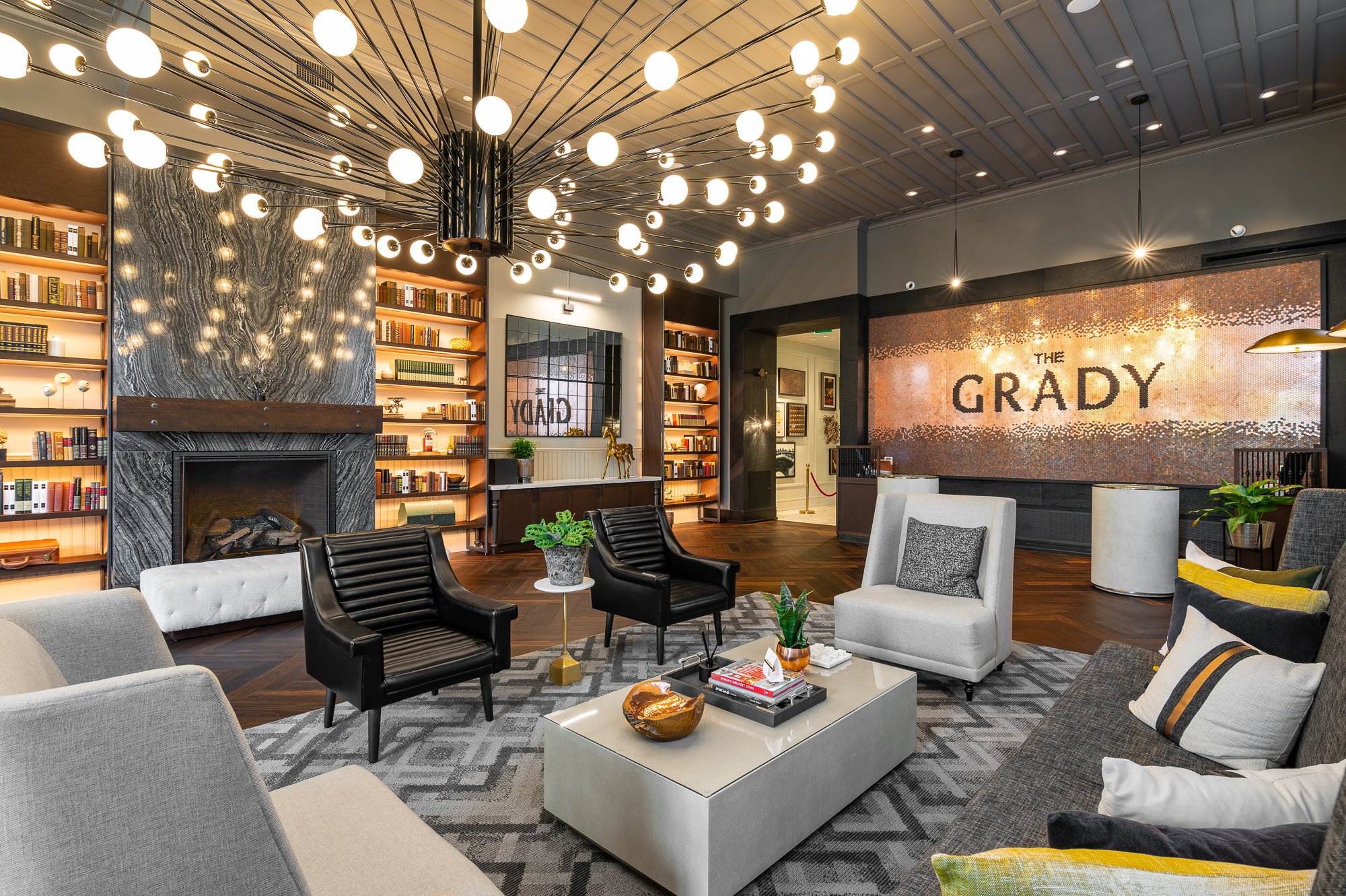 Modern hotel lobby with a large abstract fireplace, bookshelves, and seating area, illuminated by a contemporary chandelier.