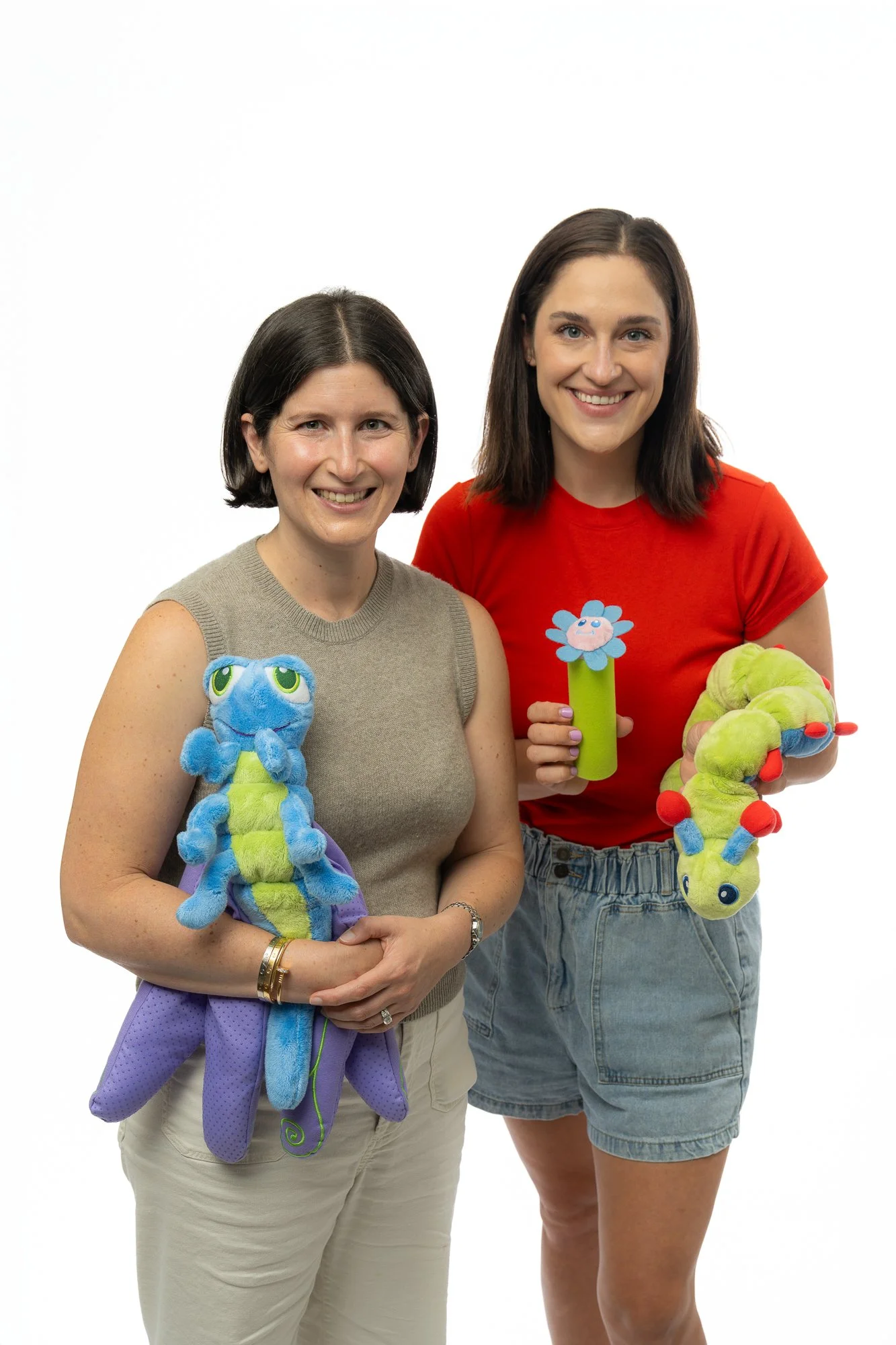 Two women standing against a white background, smiling, each holding plush caterpillars.