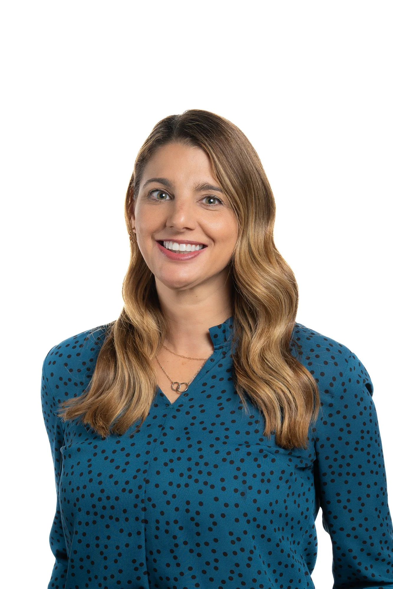 A woman with long wavy blonde hair, wearing a blue blouse with black polka dots and a layered necklace, smiling against a white background.