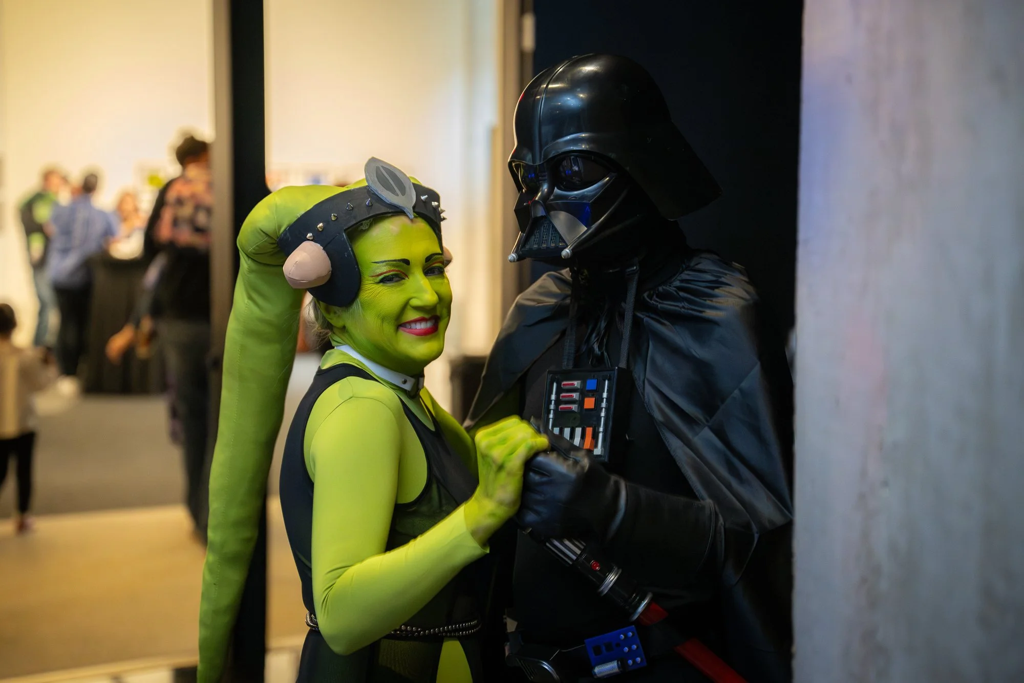A woman dressed as a green alien character from Star Wars, smiling and holding hands with a person dressed as Darth Vader from Star Wars, in a crowded indoor event.