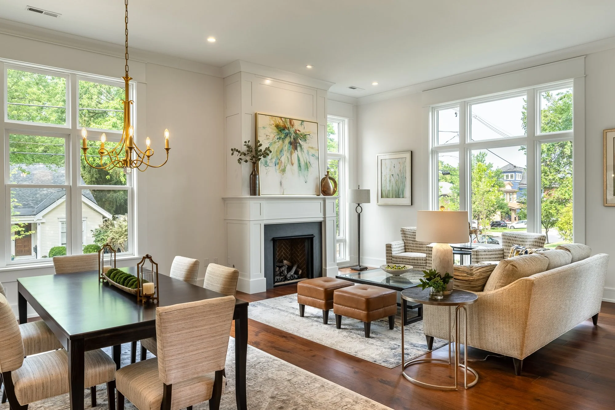 Living room with large windows, white walls, fireplace, beige sofa, upholstered chairs, wooden flooring, and modern art.