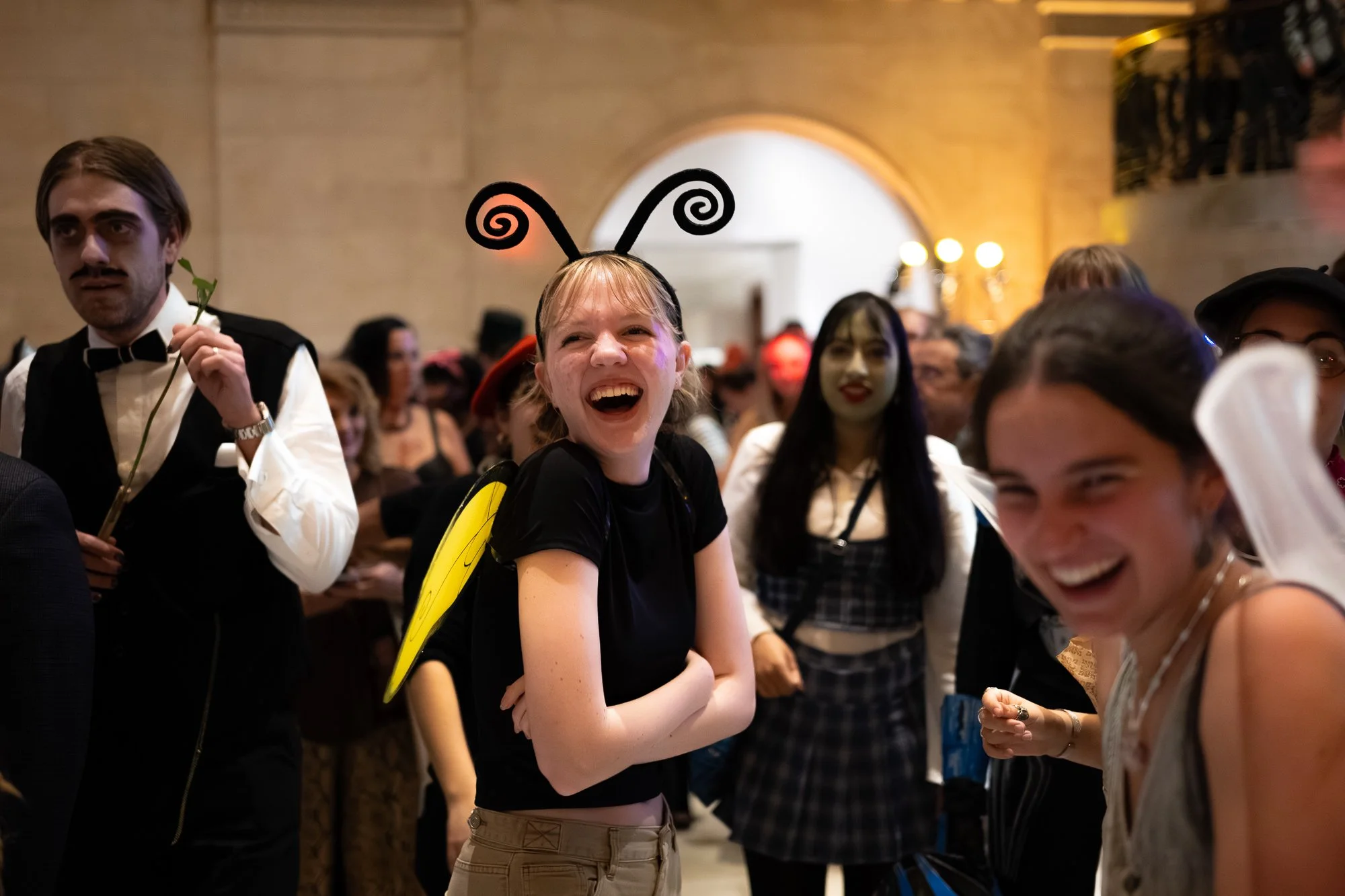A group of people at a costume party, with a woman dressed as a bee, wearing antennae and yellow wings, laughing and smiling among the crowd.