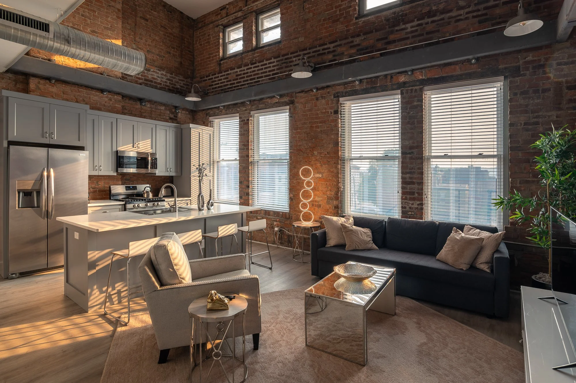 Interior view of a modern loft apartment with an open kitchen and living room. The kitchen features stainless steel appliances, gray cabinets, and an island with bar stools. The living area has a dark sofa with pillows, a glass coffee table, and a be