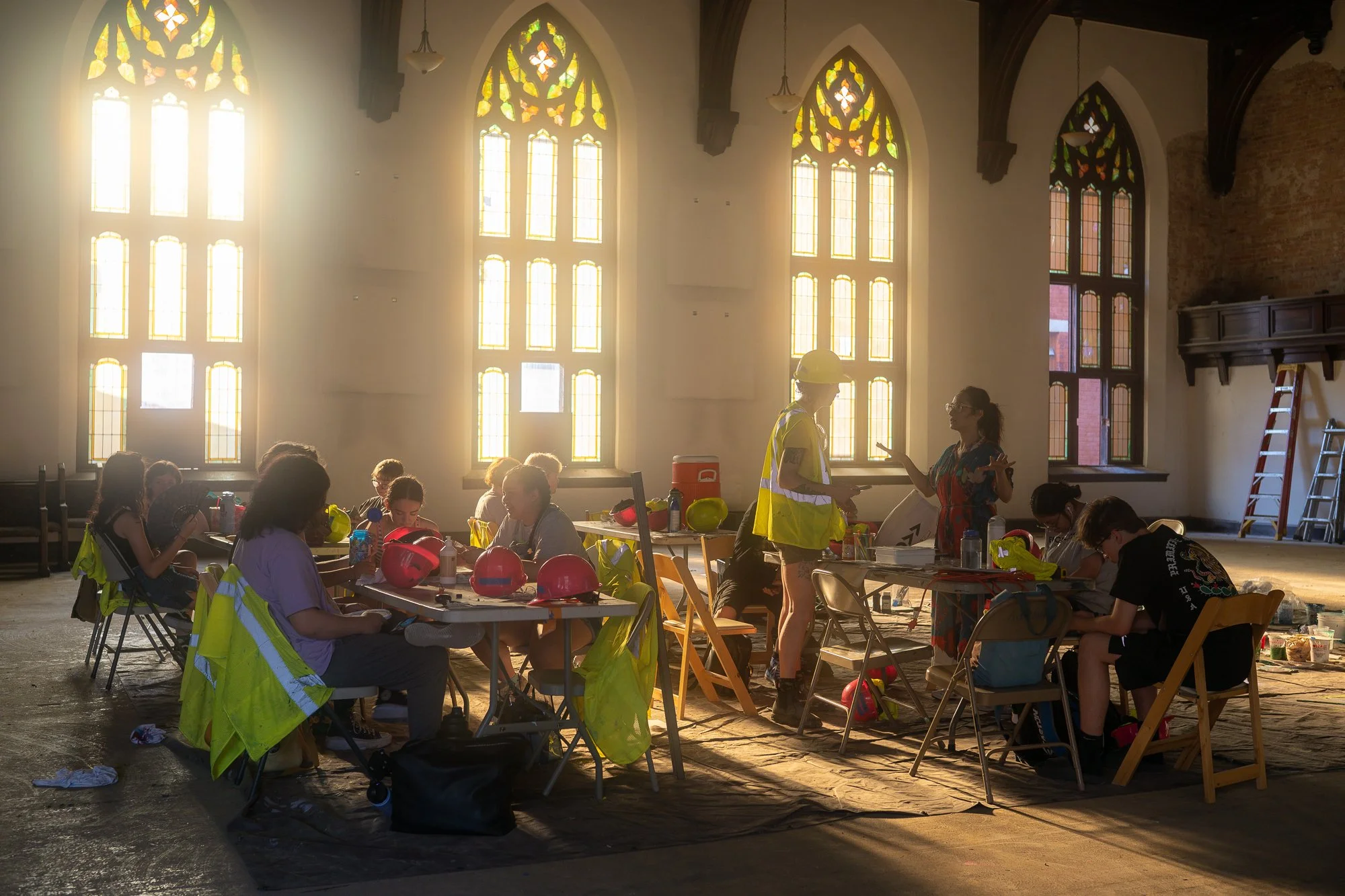Group of people wearing reflective vests and helmets sitting at tables inside a large room with tall stained glass windows, some people working or socializing, with construction equipment and ladders in the background.
