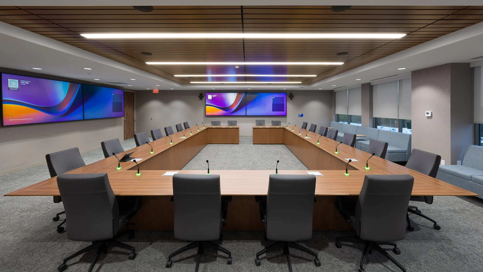 Empty conference room with U-shaped wooden table, black chairs, microphones, and large digital screens on the wall.