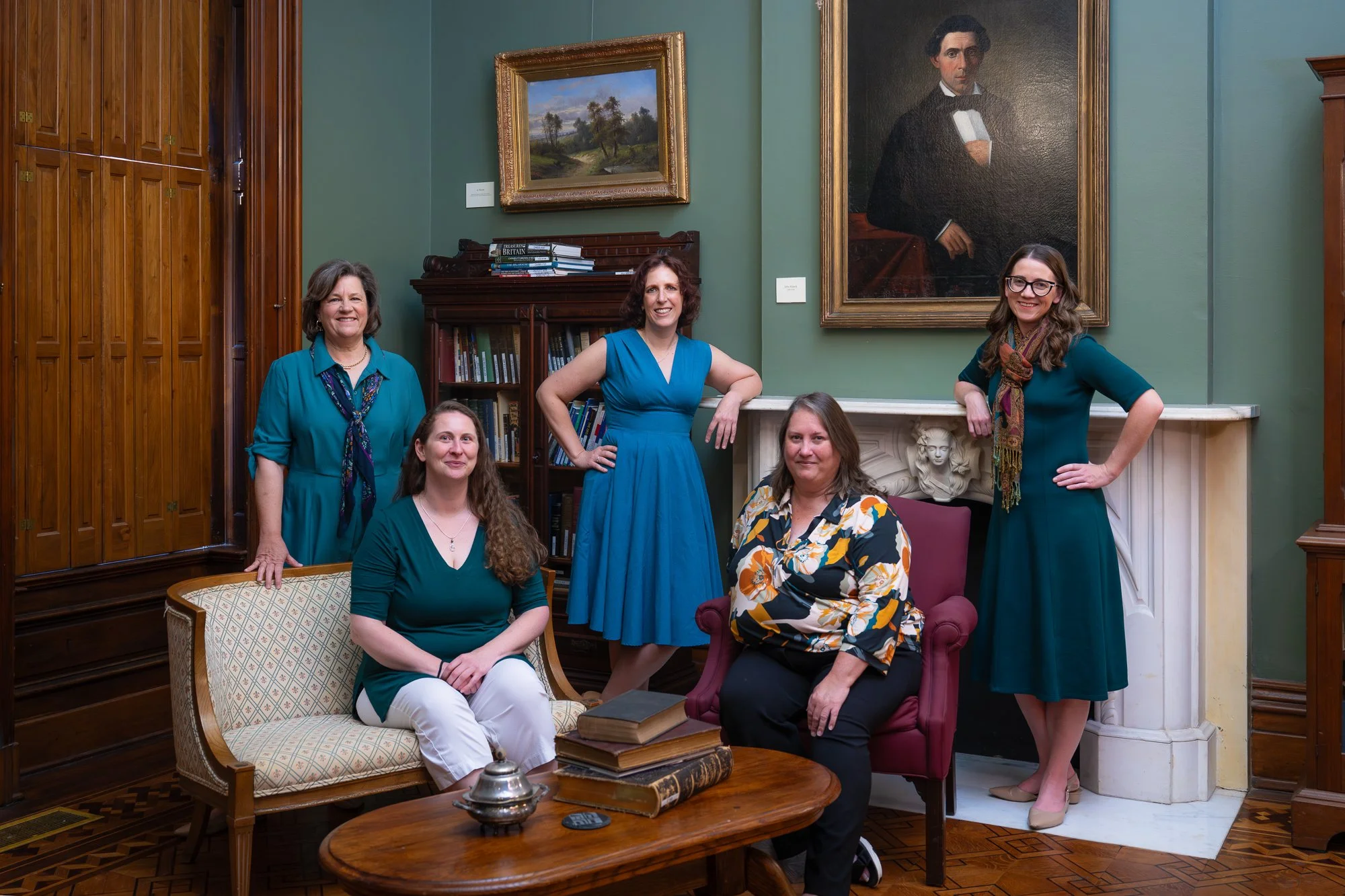 Five women in a room with green walls and classical decor, sitting and standing around a fireplace and a bookshelf, with portraits hanging on the wall behind them.