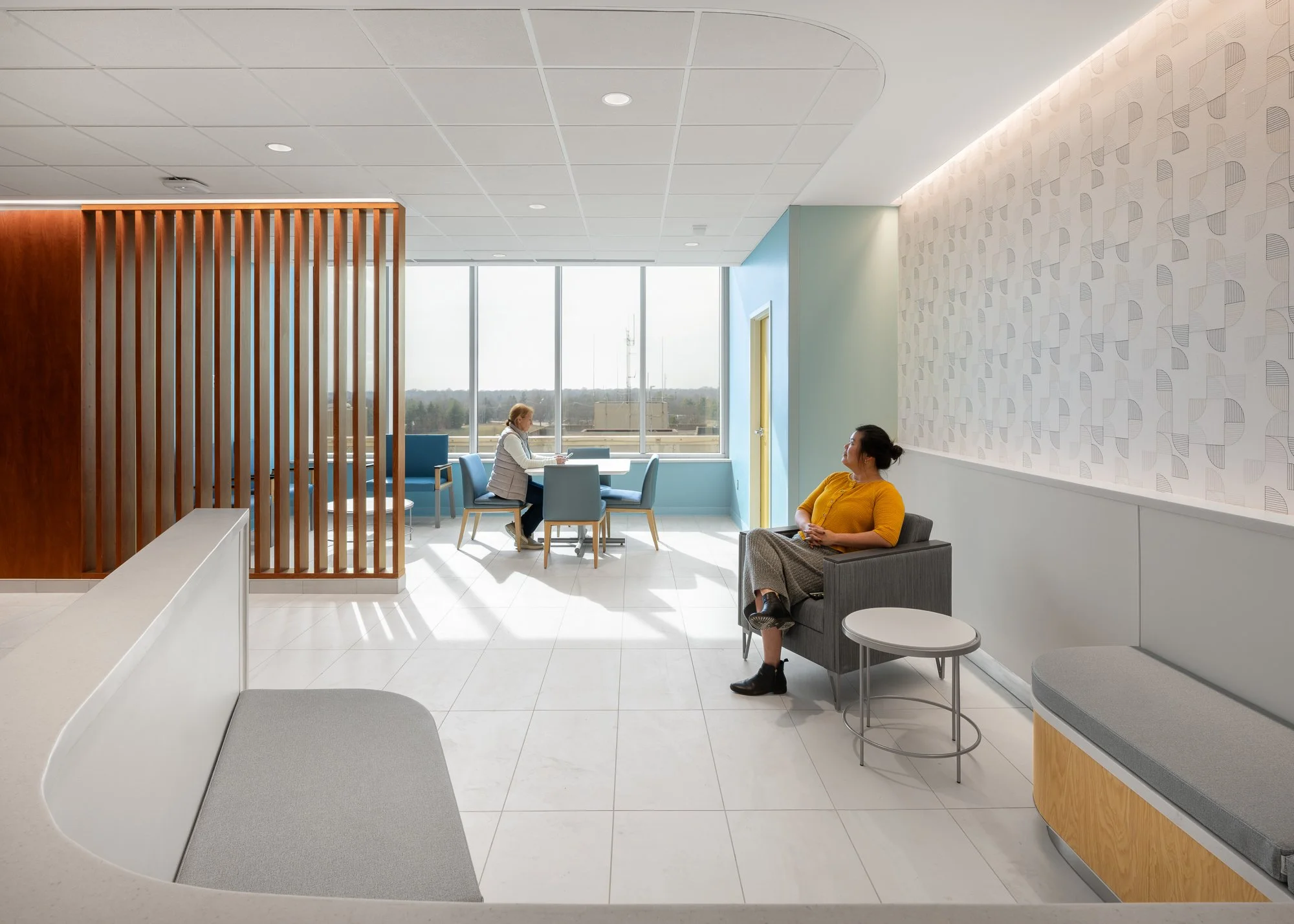 A modern medical waiting room with two women seated, one in a wheelchair and the other at a table, with large windows and patterned wall decor.