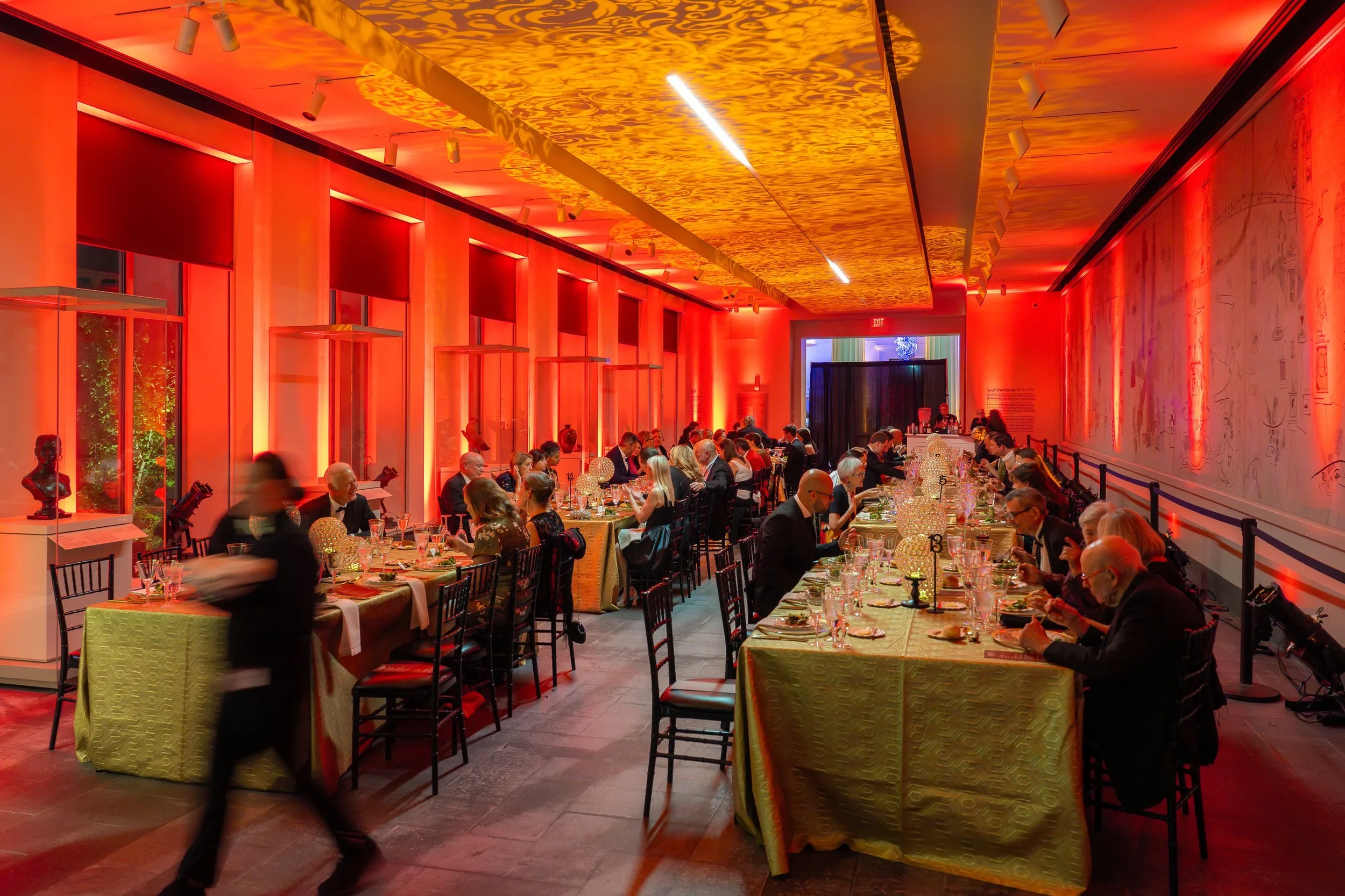 An elegant indoor banquet hall decorated with warm orange and gold lighting, filled with round and rectangular tables set for a formal dinner with guests dressed in formal attire, and a waiter carrying a tray walking through the room.