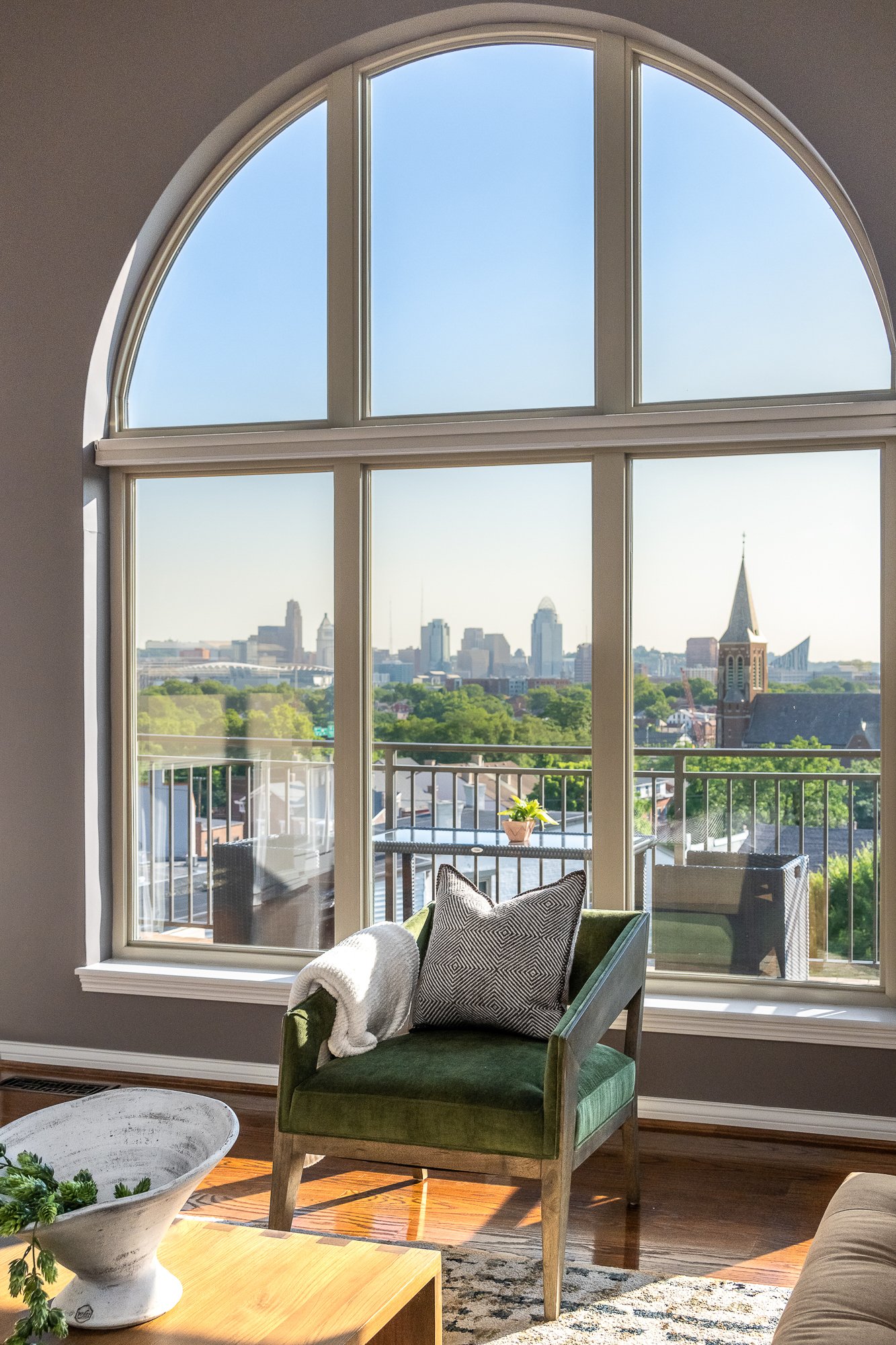Living room with large arched window overlooking a city skyline, with a green armchair, pillows, a blanket, and a small plant on a balcony.