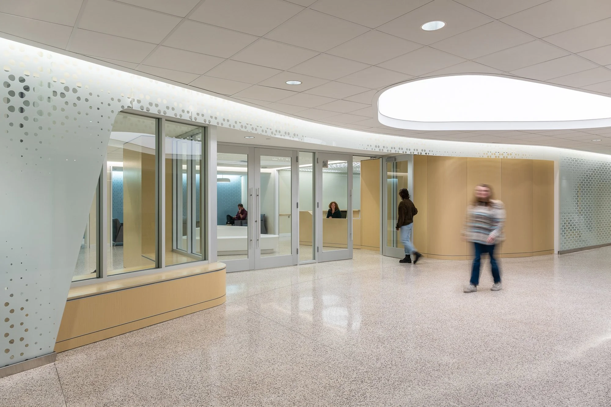Modern hospital or medical office lobby with glass doors, reception desk, and seating area, with three people walking or sitting inside.