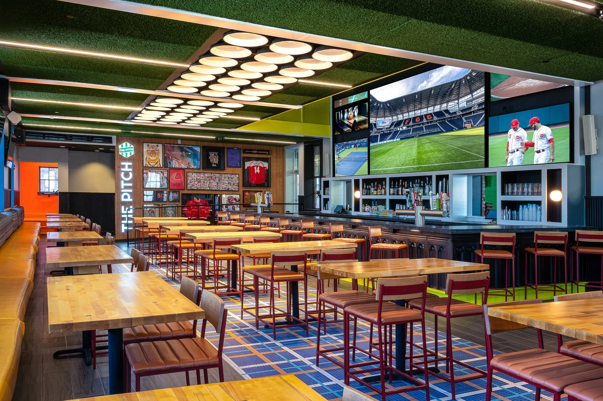 A sports bar with multiple large screens showing a baseball game and a stadium field, wooden tables and chairs, a bar counter with bottles, sports memorabilia on the wall, and a ceiling with modern lighting fixtures.