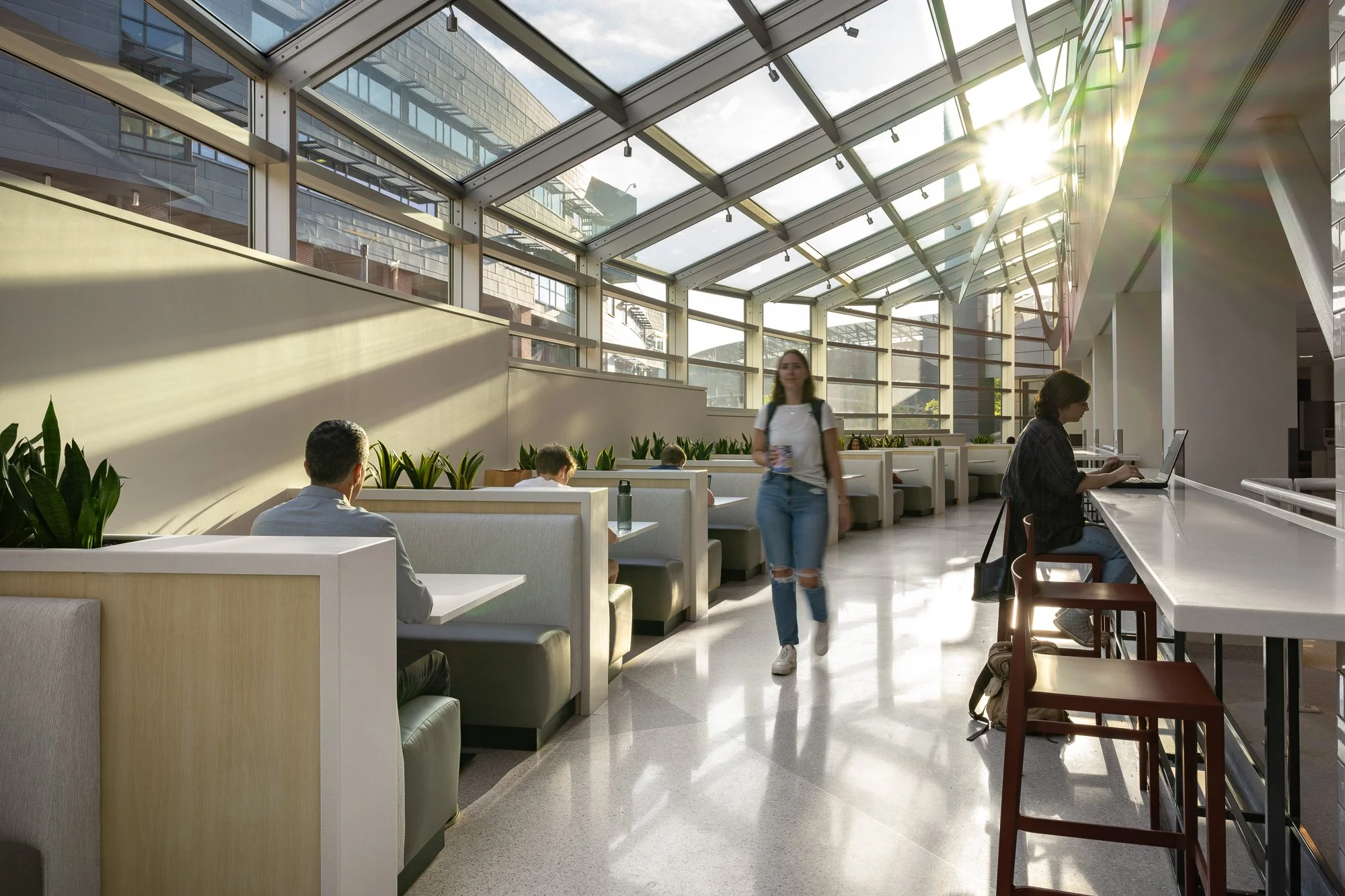Sunlit modern cafe or food court with booths and bar seating, glass roof, and people working or sitting