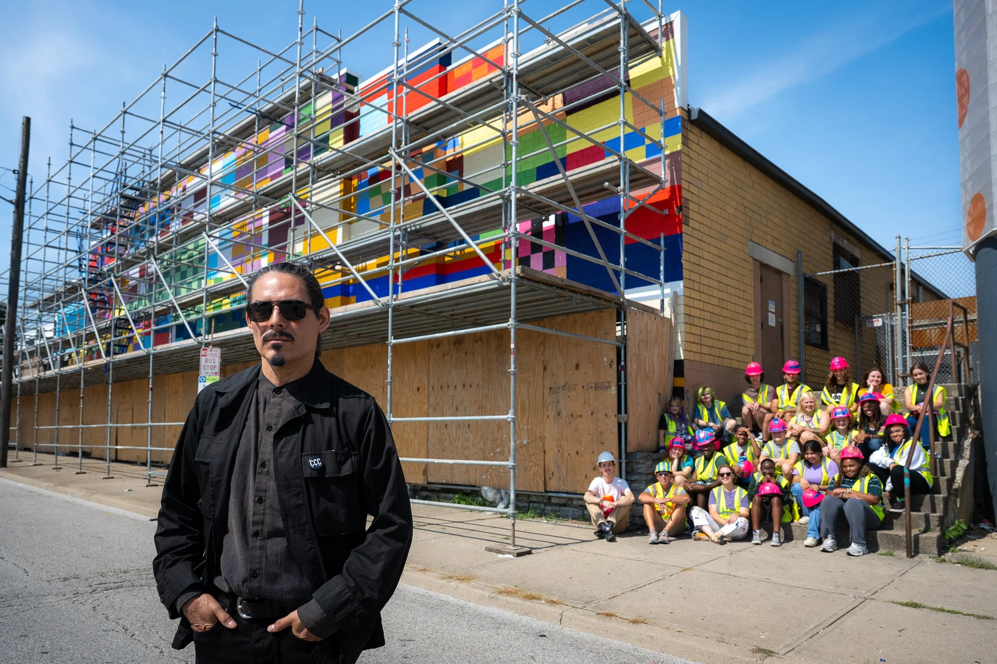 A man in black clothing and sunglasses stands in front of a construction site with colorful murals and a group of children and adults in safety vests and helmets sitting on stairs.
