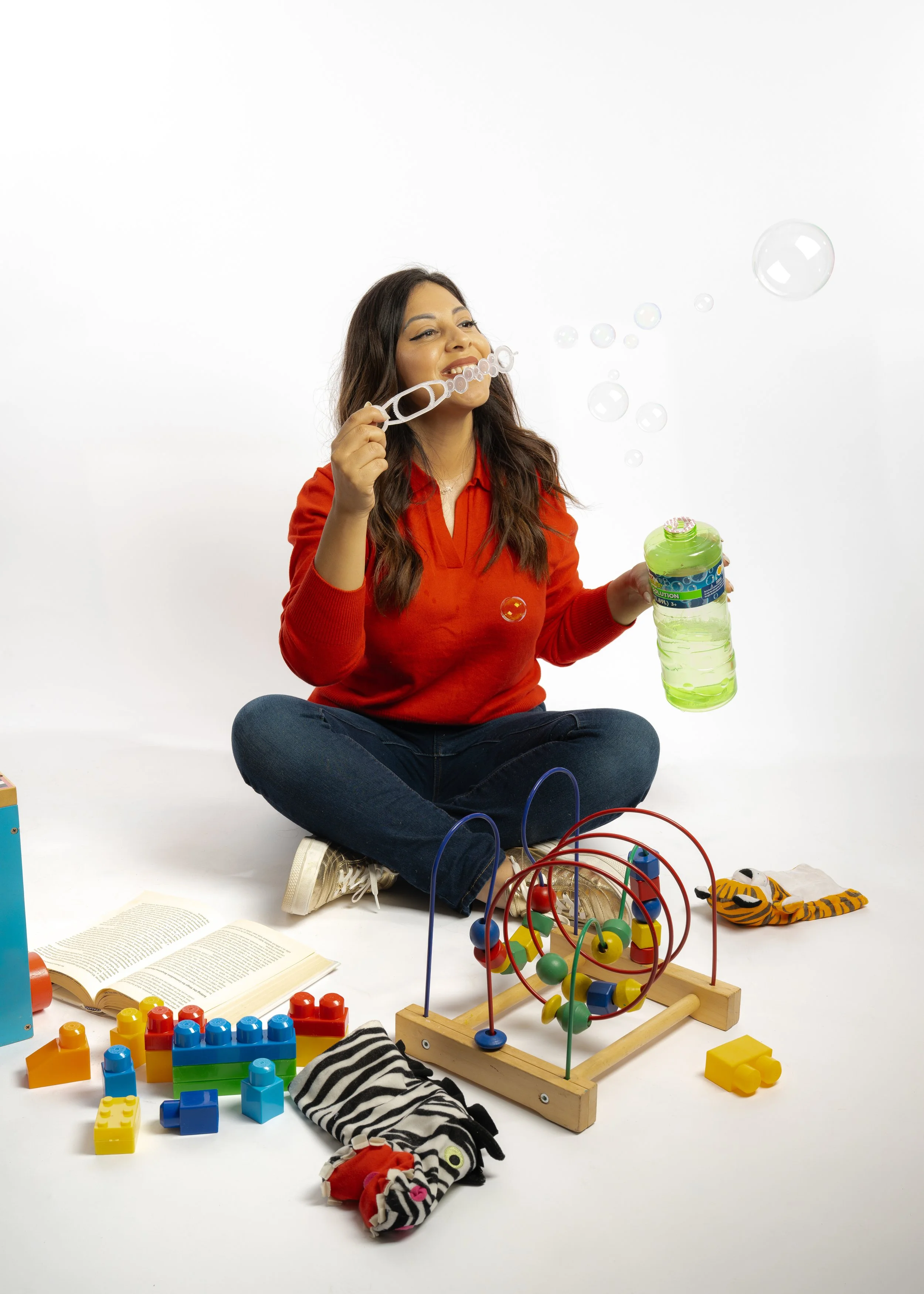 Woman sitting cross-legged on the floor blowing bubbles with a bubble wand, surrounded by toys including a bead maze, toy blocks, a plush zebra, and a book, against a white background.