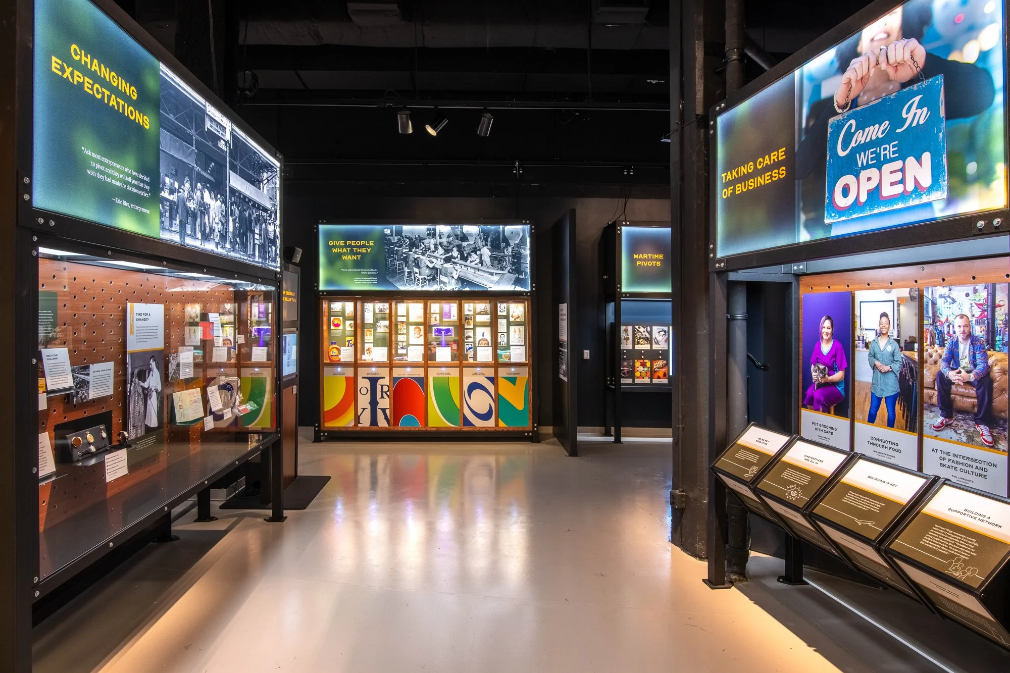 An interior of a museum exhibit with digital displays and informational panels about social change, community, and history. Bright screens display quotes, images, and photos of people and activities.