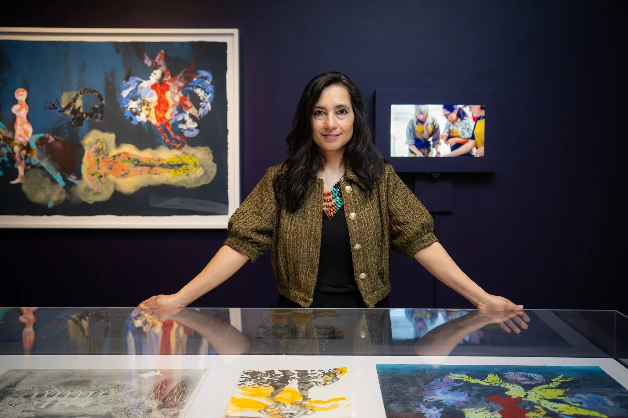 A woman with long dark hair standing behind a glass display case in an art gallery, with colorful artwork behind her and a small video screen on the wall.
