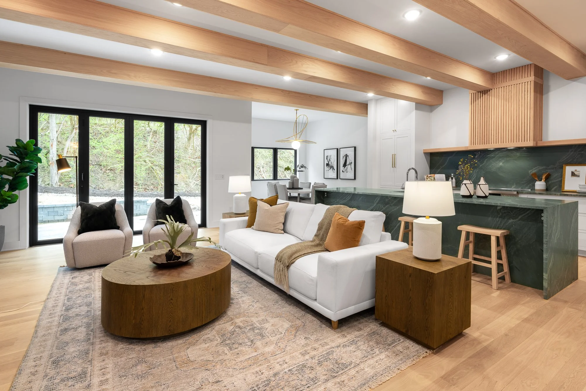 Modern living room with white sofa, gray armchairs, wooden coffee table, and large sliding glass doors leading to outdoor greenery. Open-concept kitchen with green marble counter, white cabinets, and dining area with pendant light.