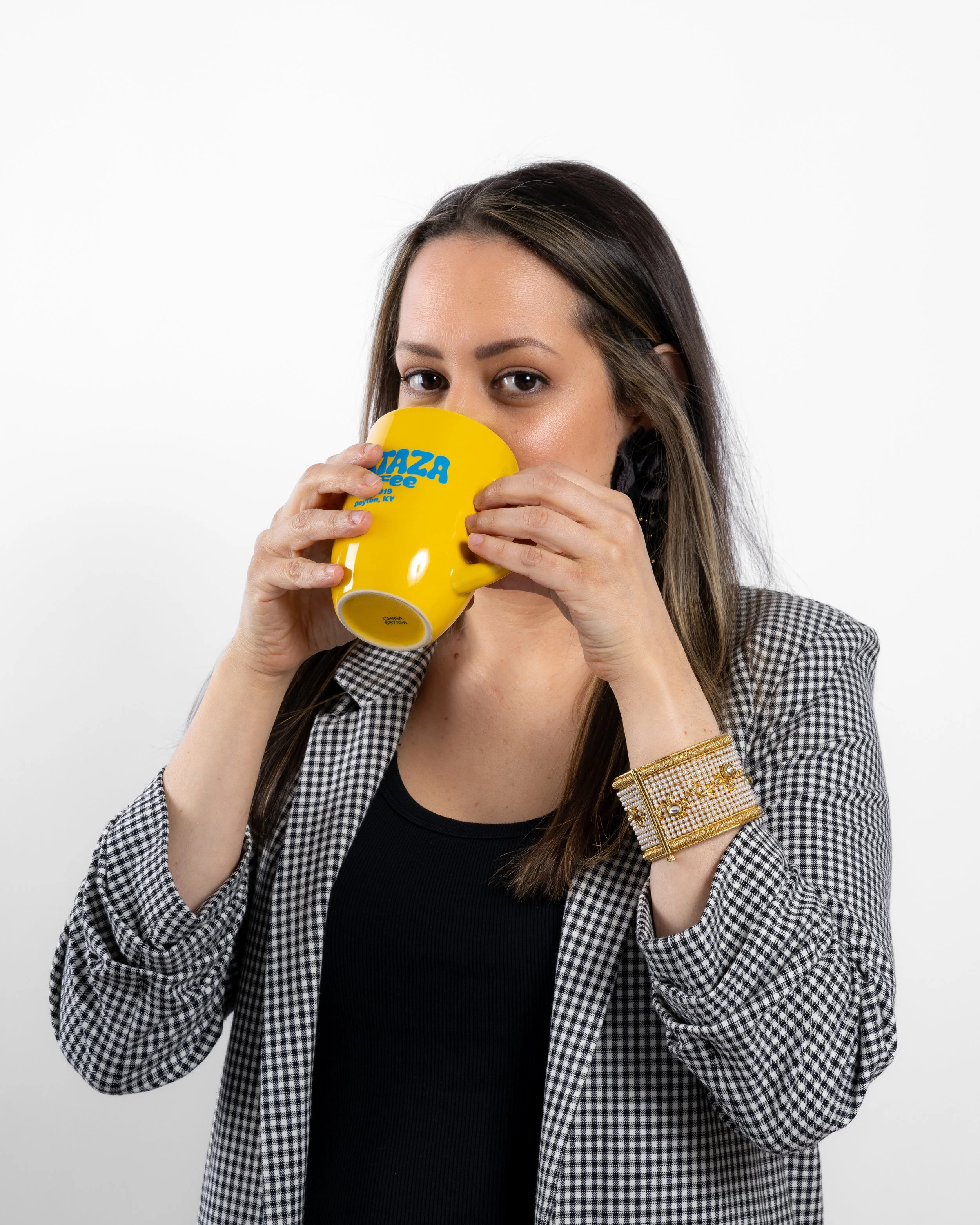 Woman with long dark hair wearing a checkered blazer and black shirt, holding a yellow mug with blue text, drinking from it, standing against a plain white background.