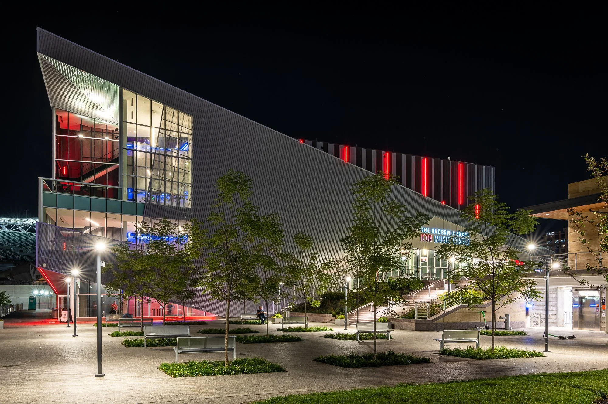 Night view of a modern building with illuminated windows, colorful lights, and a landscaped walkway with trees and benches in front.
