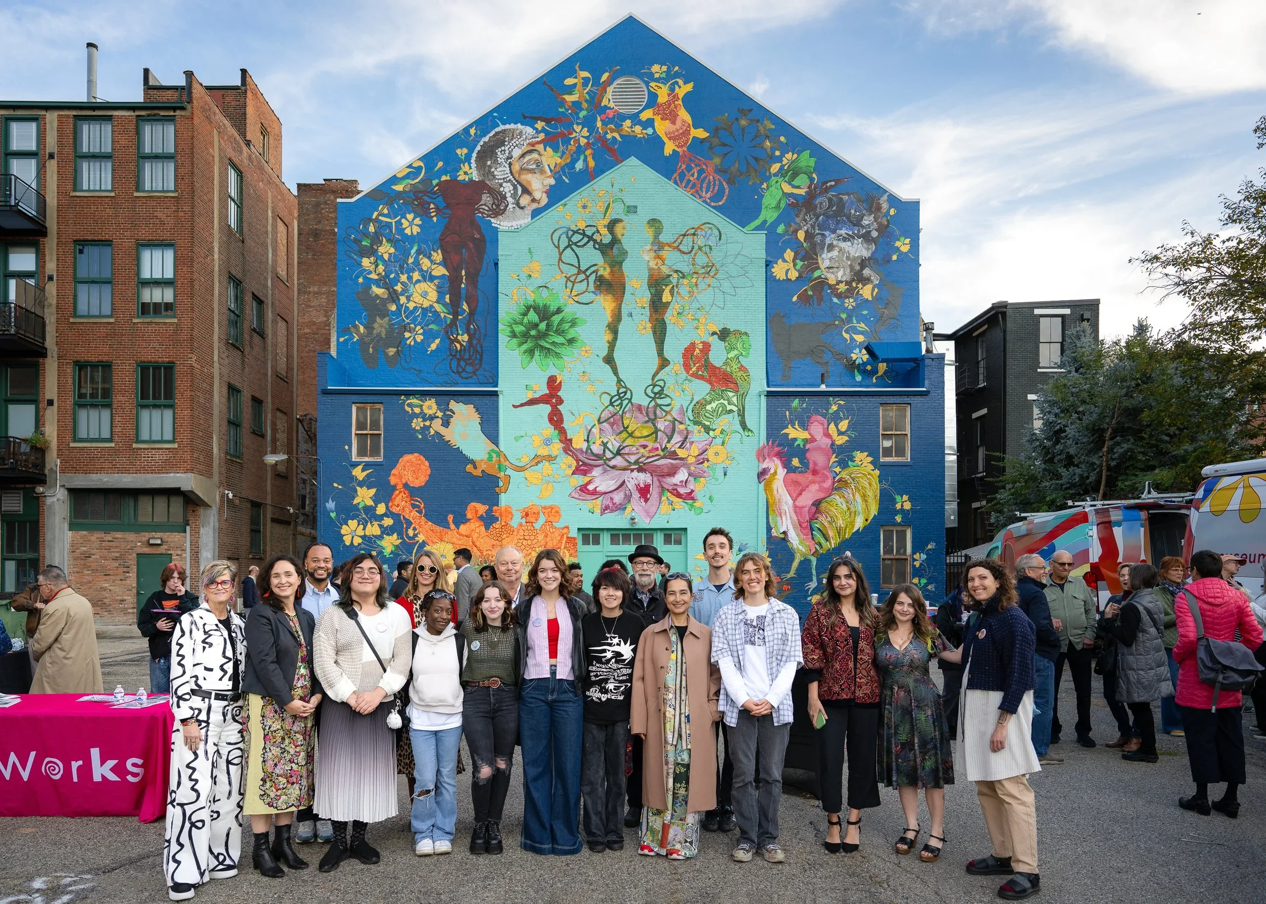 A group of people standing in front of a large colorful mural on a city street, with buildings and a sky in the background.