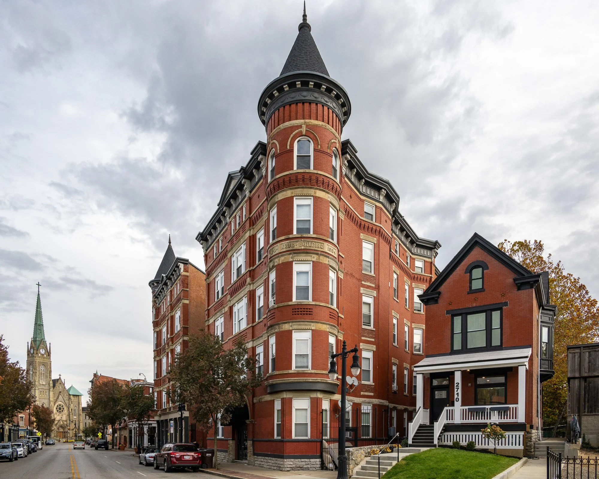 A tall, historic red brick building with a rounded tower topped by a pointed spire. The building has multiple windows and decorative architectural details. There are smaller houses and a church steeple visible in the background on a cloudy day.