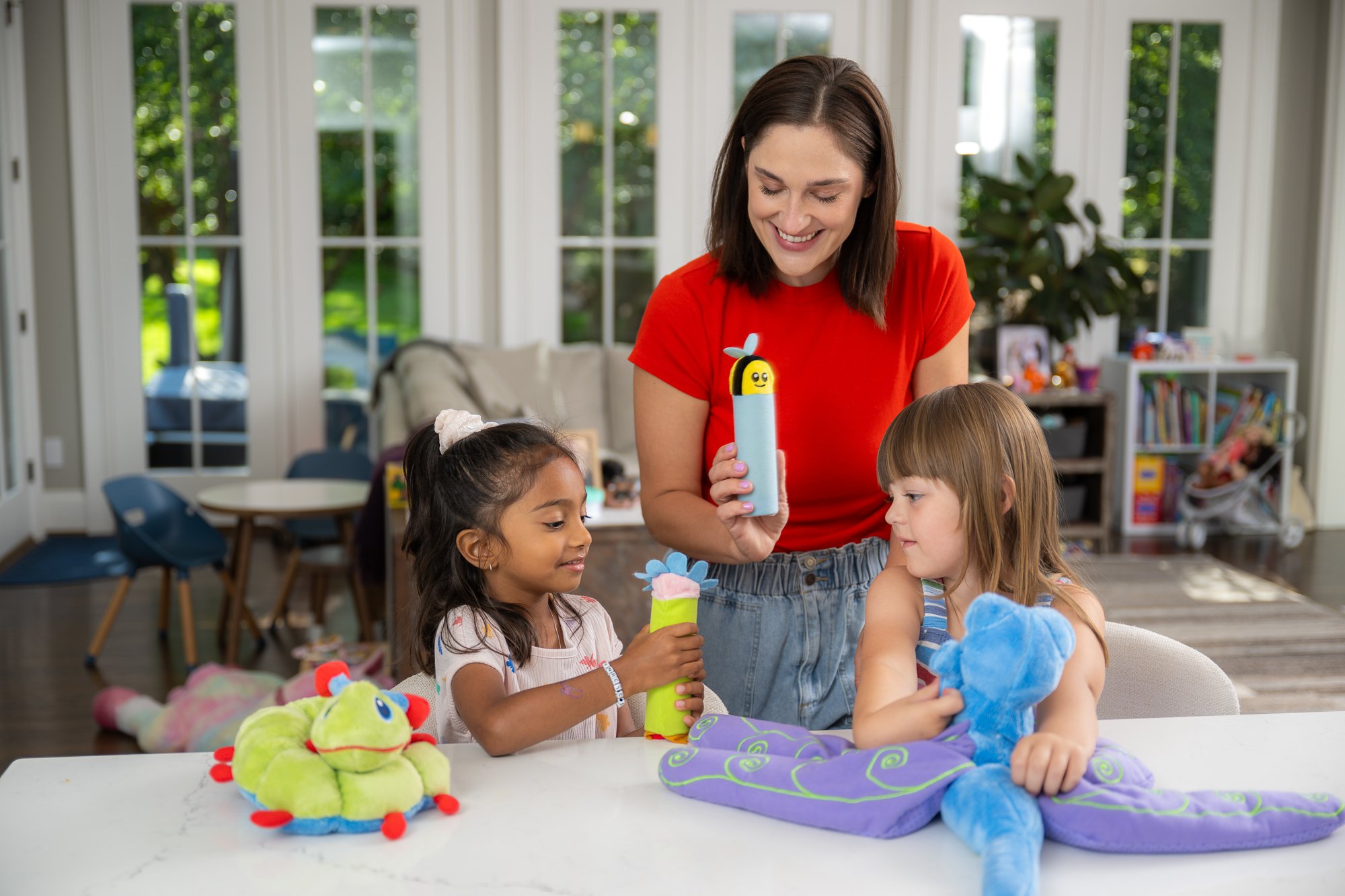 A woman and two young girls playing with plush toys and cylindrical objects decorated as bees and frogs inside a bright living room with large windows and greenery outside.