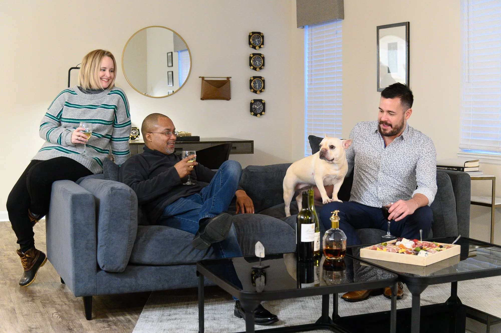 Four people and a dog in a living room. Three of them are sitting on a gray sofa, and one woman is leaning over it, holding a glass of wine. The man on the right is sitting on the armrest, also holding a glass of wine, with a small dog standing on th