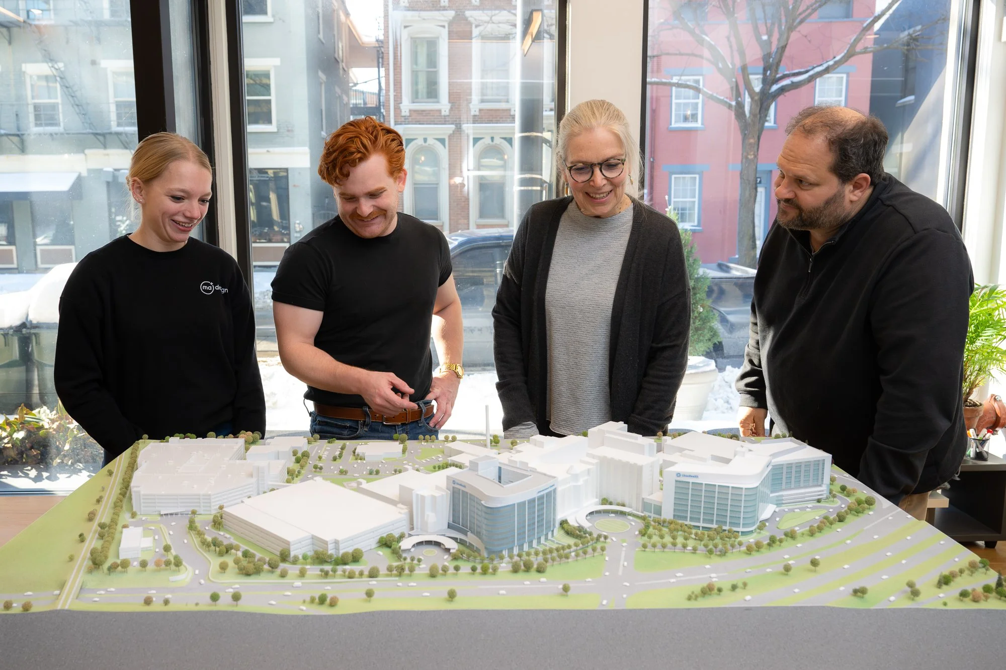 Four people looking at a scale model of a building development project indoors with large windows in the background.