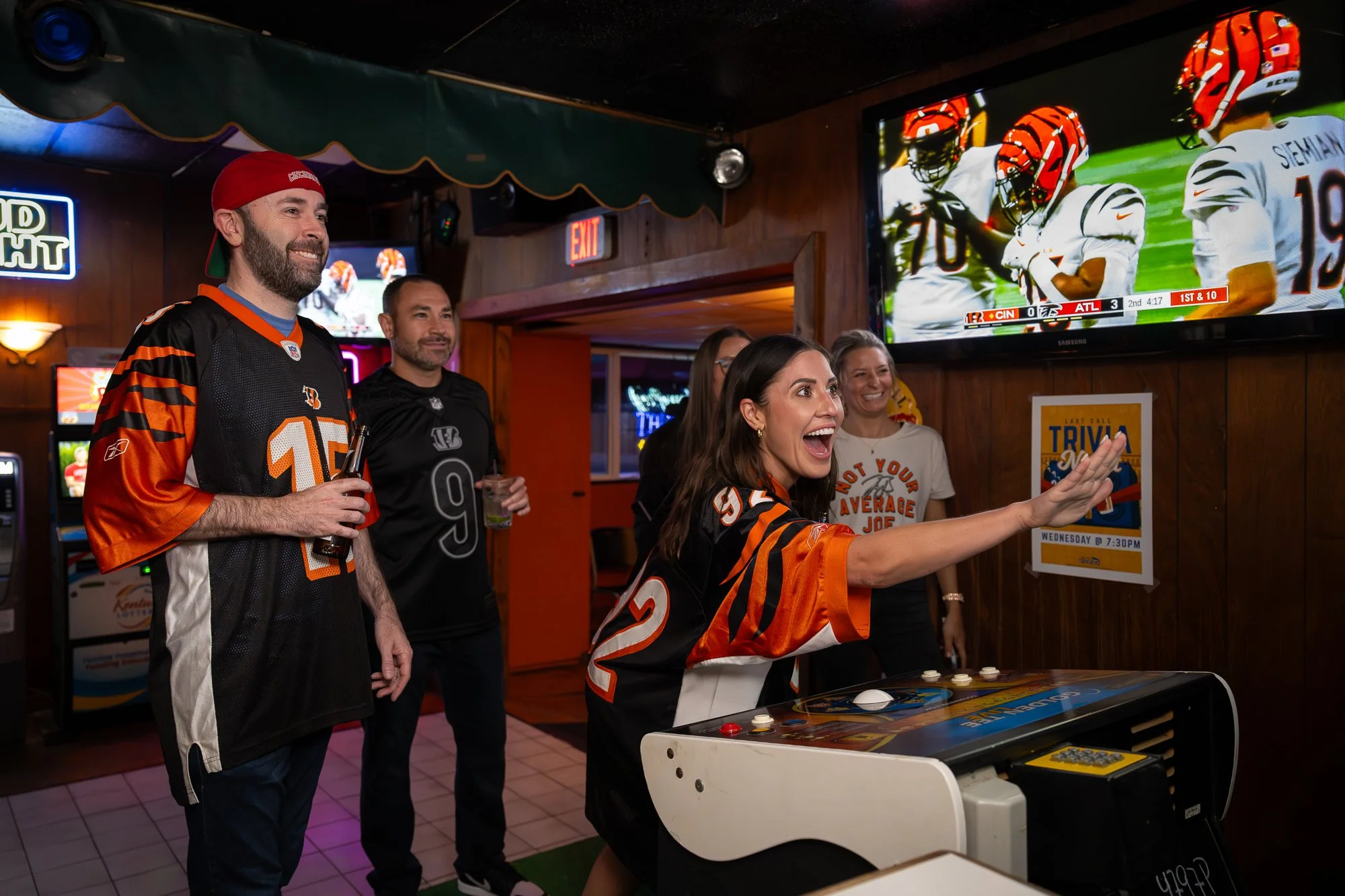 People at a bar watching a football game on TV, with some wearing Cincinnati Bengals jerseys, and one woman playing a video game machine.