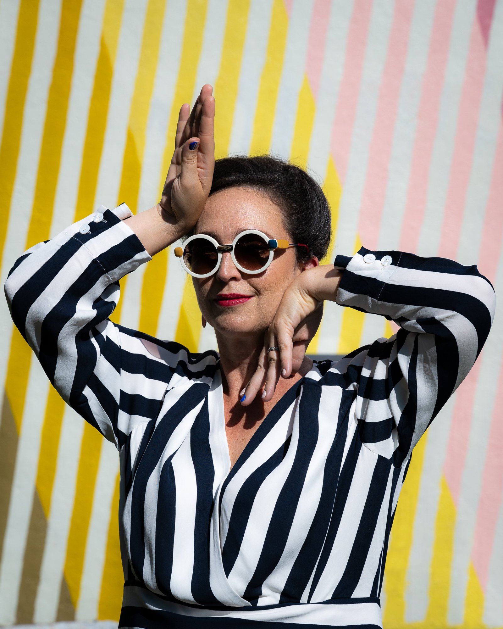 A woman with dark hair wearing large, round sunglasses and a black and white striped shirt, posing with her hand on her head and her other hand near her chin, in front of a colorful striped background.