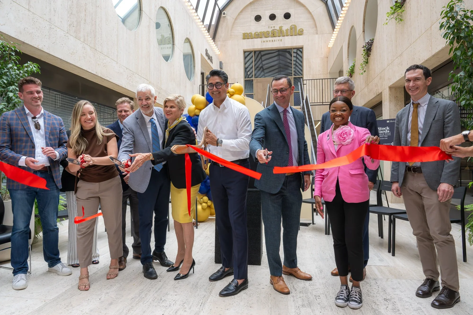 Group of people at ribbon-cutting ceremony inside a modern building with balloons and cardboard sign that says "The Mercantile".