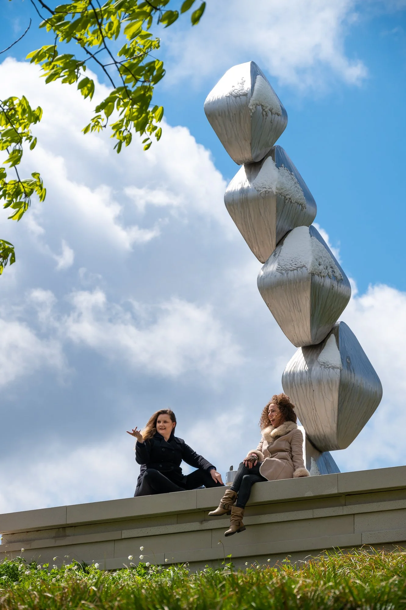 Two women sitting on a ledge near a modern, abstract sculpture made of large, shiny, metallic, geometric shapes. One woman is gesturing, and the other is smiling. The scene is outdoors with a partly cloudy sky and green foliage.