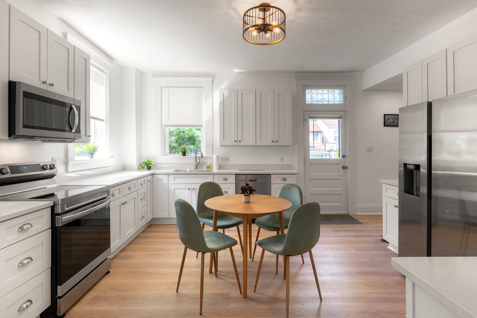 Bright kitchen with white cabinets, stainless steel appliances, a round wooden dining table with four green chairs, and a window overlooking greenery.