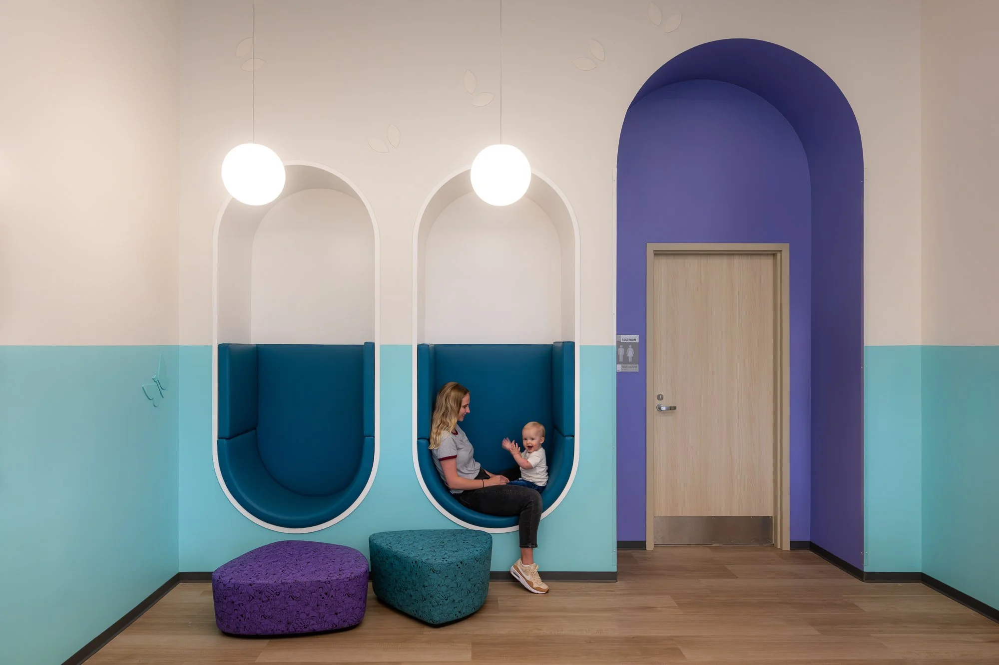 A woman and a baby sitting in a colorful modern waiting area with two blue pod chairs, purple and teal ottomans, and a purple accent wall.