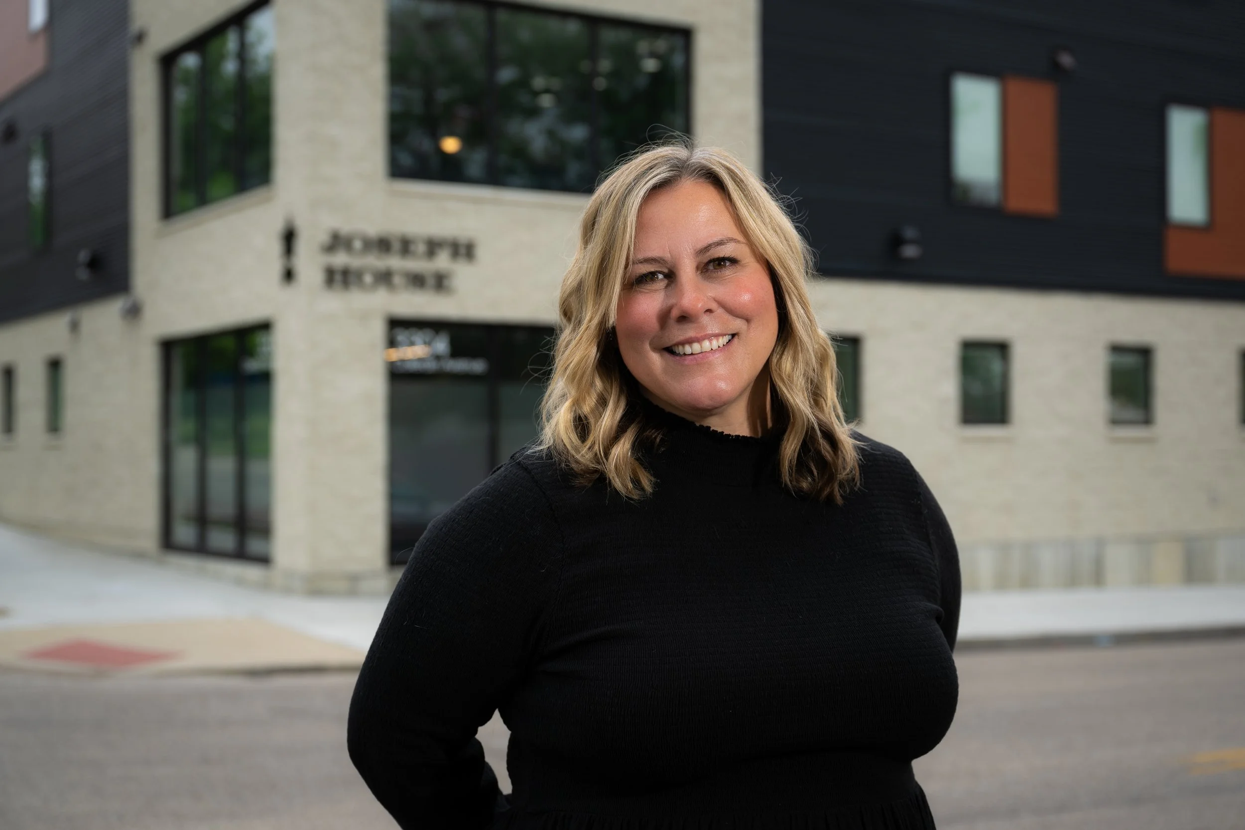 A woman with blonde hair wearing a black sweater standing outside in front of a modern building with a sign that reads 'Joseph House'.